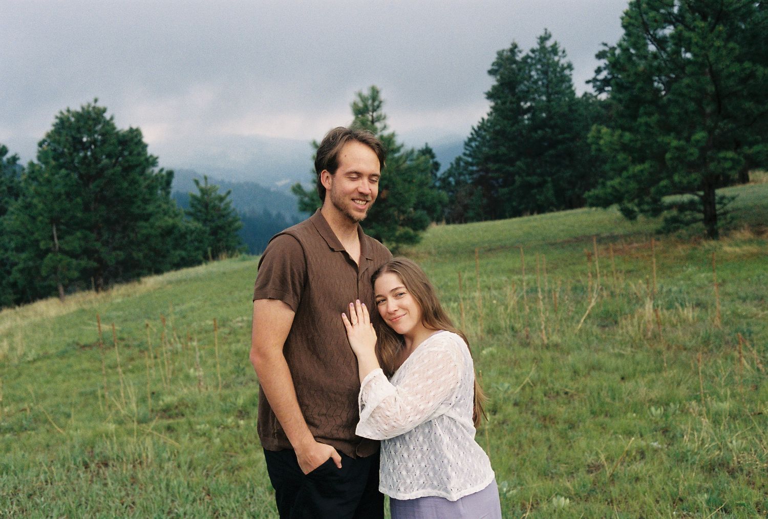 A smiling couple embraces in a lush green meadow with misty mountains in the background.