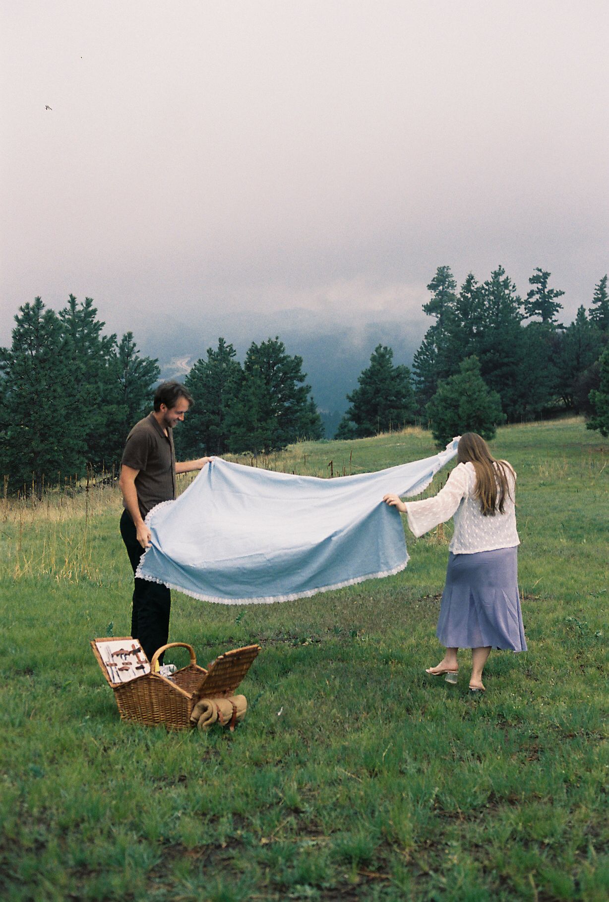 Couple spreading a light blue blanket on green meadow for a picnic, with wicker basket and dog nearby.