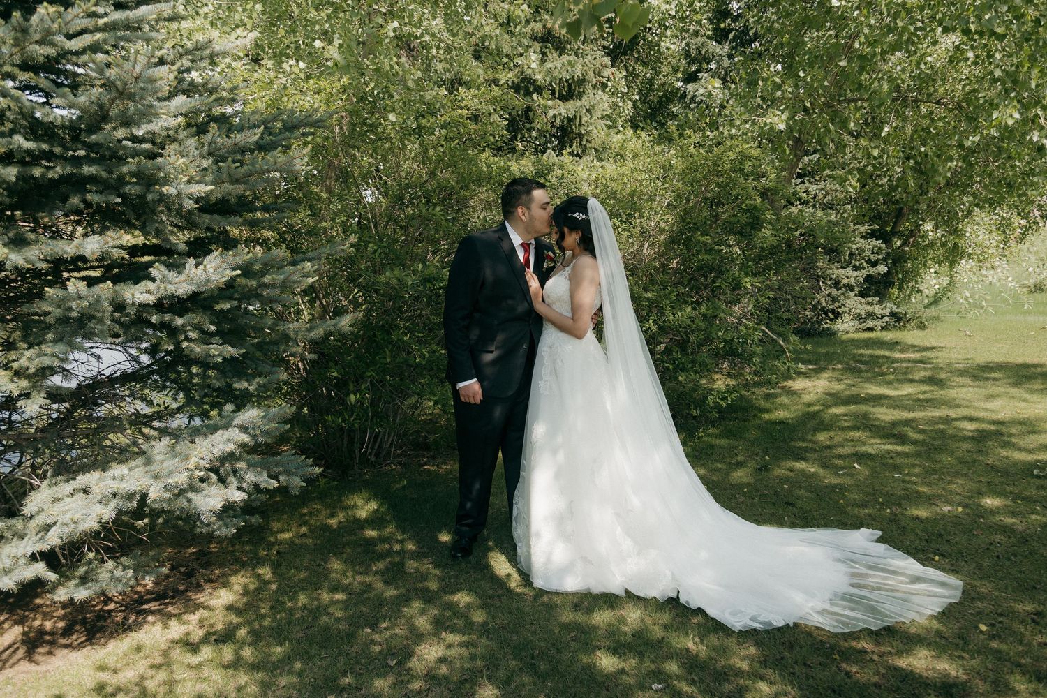 Bride and groom sharing a romantic moment among green trees with long wedding veil trailing.