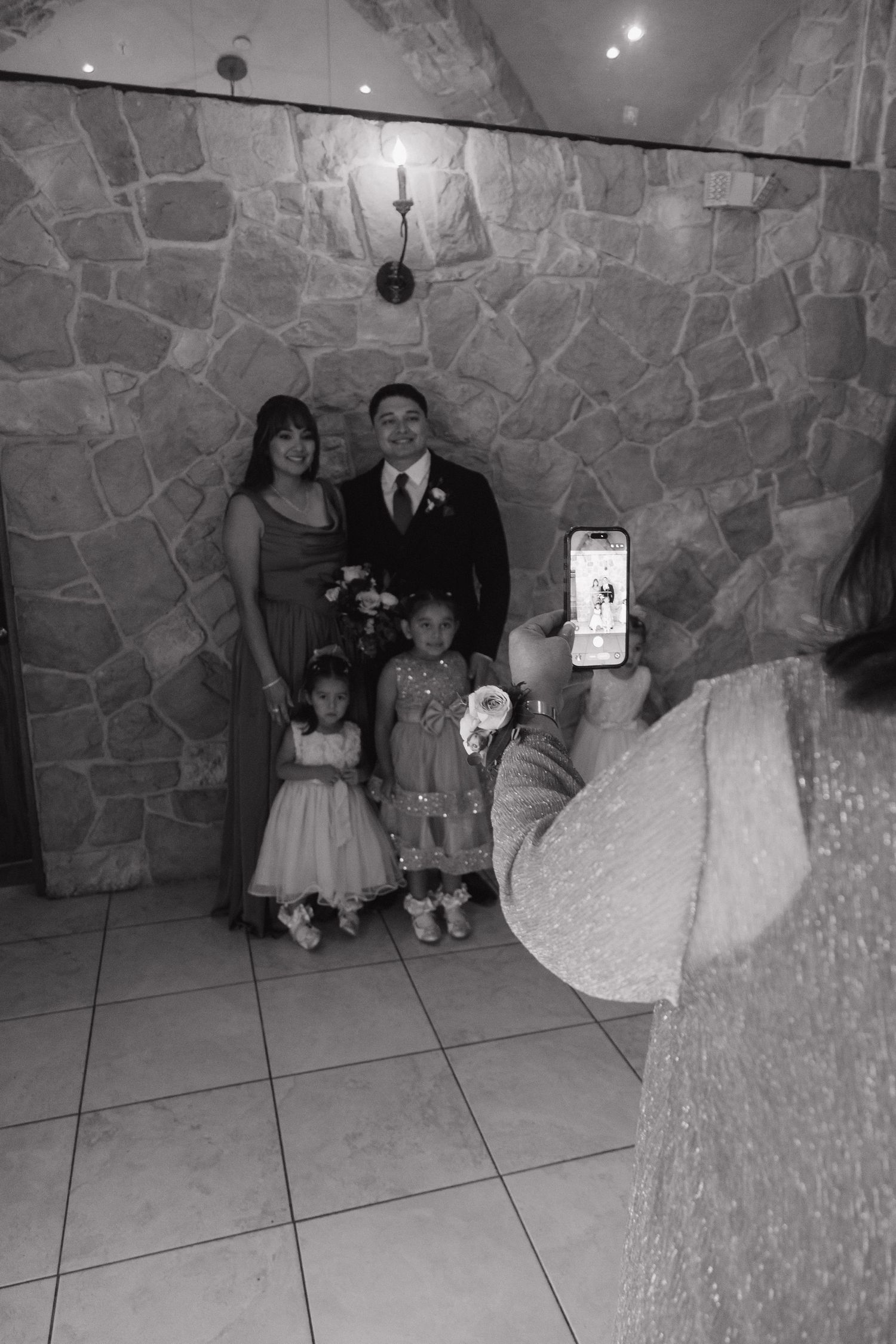 Groom poses with family and flower girls as guest photographs them at wedding venue.