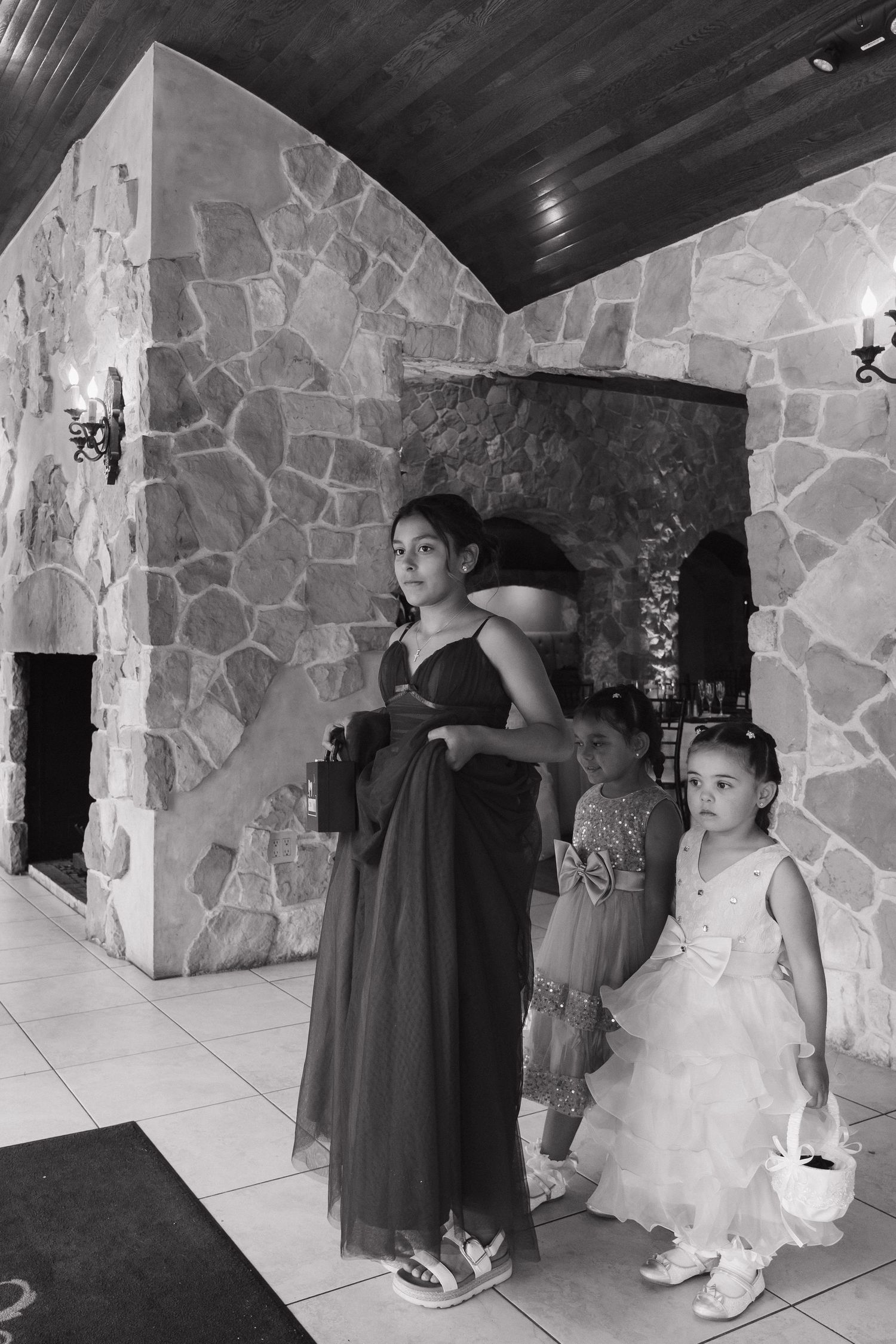 Bridesmaid in dark gown stands with two young flower girls in white dress inside stone-walled wedding venue.