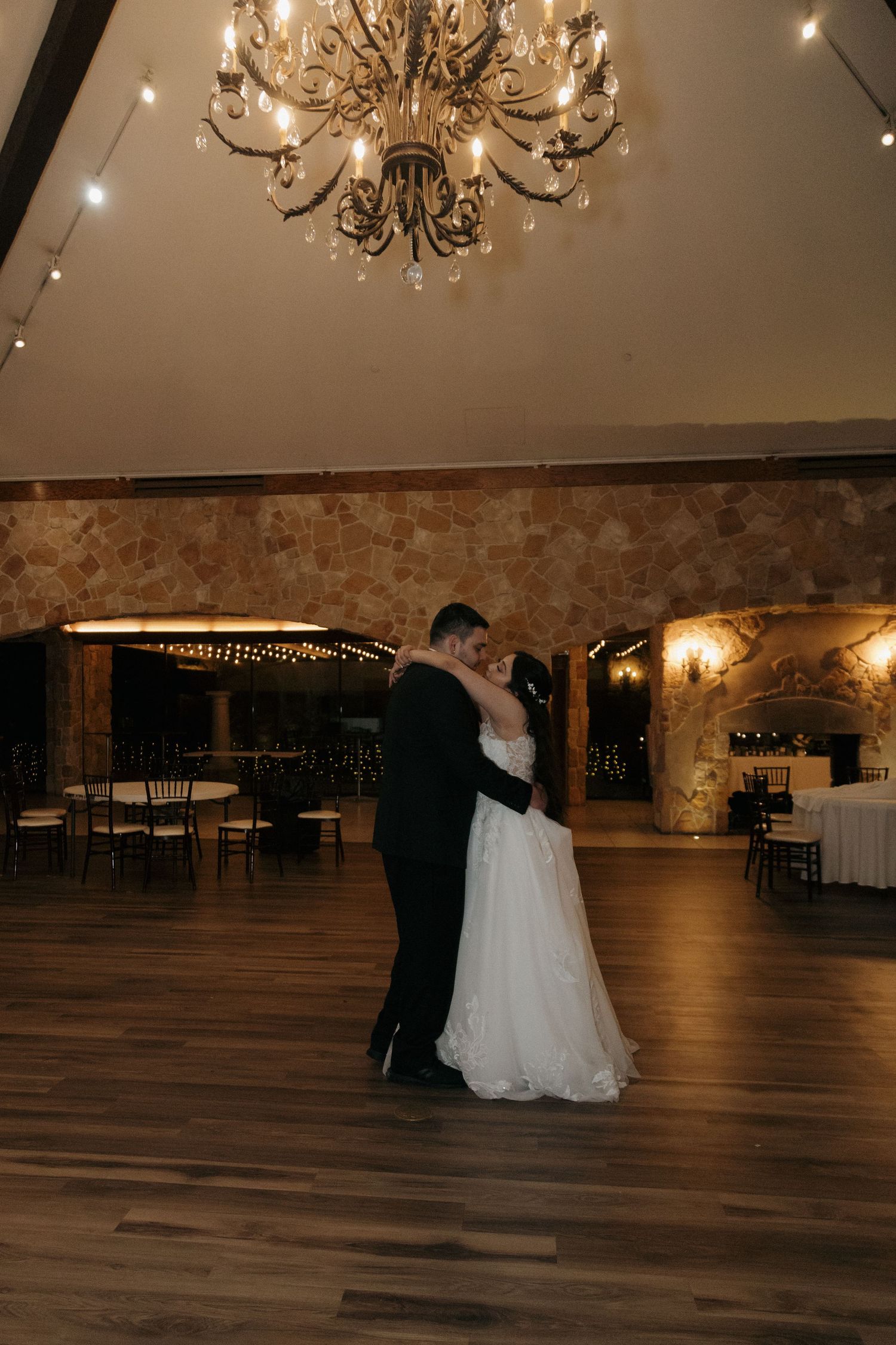 Bride and groom sharing intimate first dance under ornate chandelier in elegant stone ballroom.