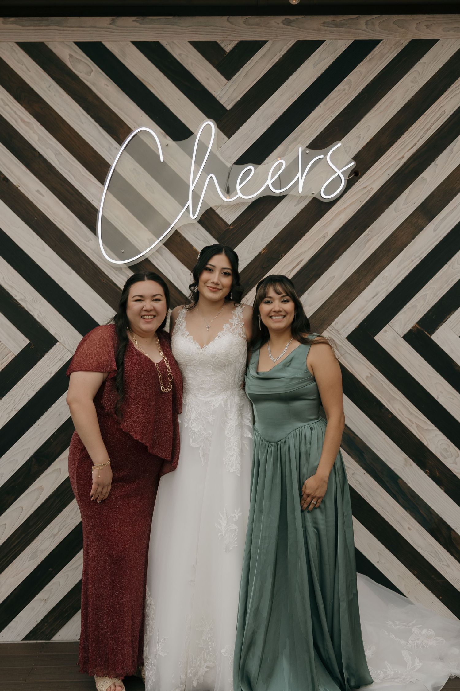 Bride in lace gown poses with two guests in front of a Cheers neon sign at wedding reception.