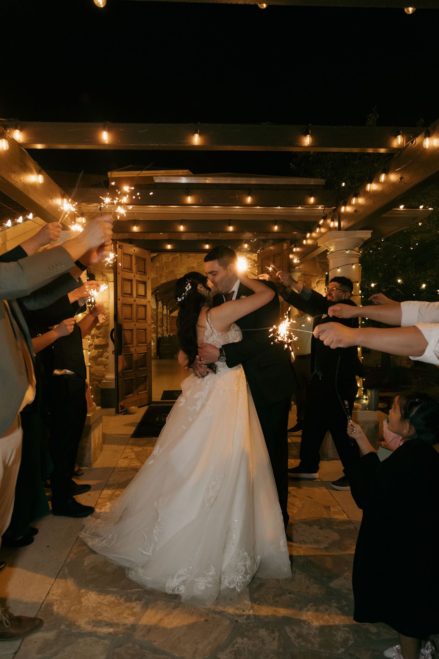 Bride and groom kiss while guests celebrate with sparklers during a romantic nighttime wedding send-off.