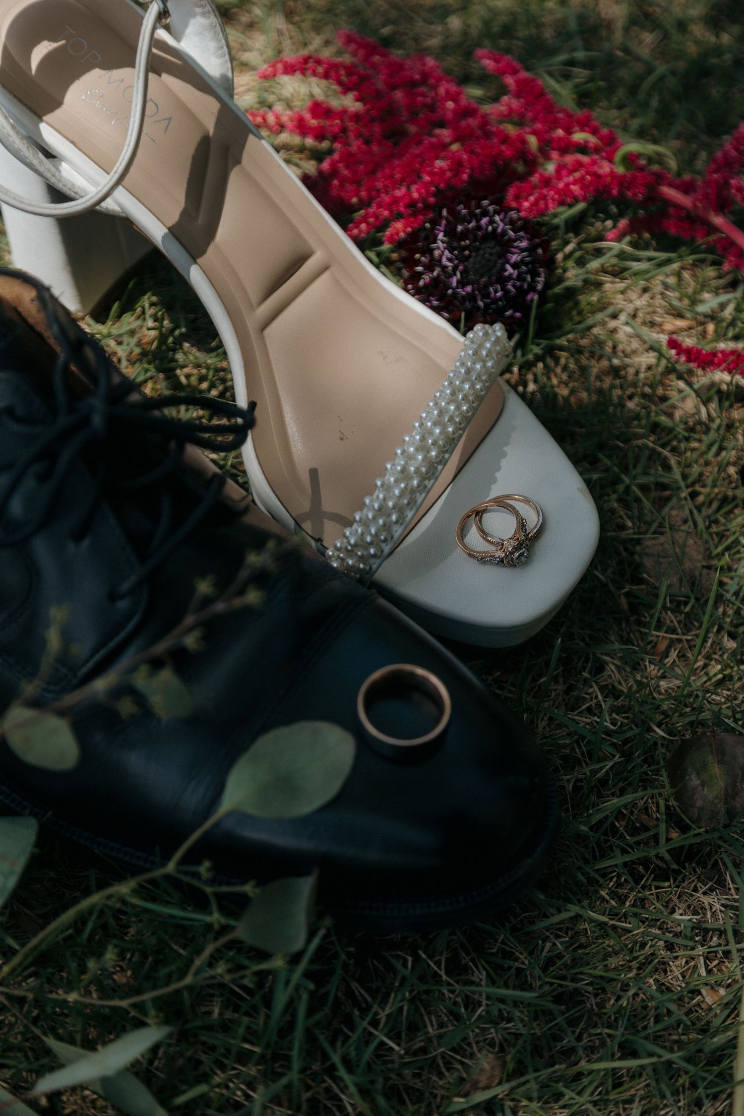White wedding heels and black dress shoes on grass with gold rings and red flowers.