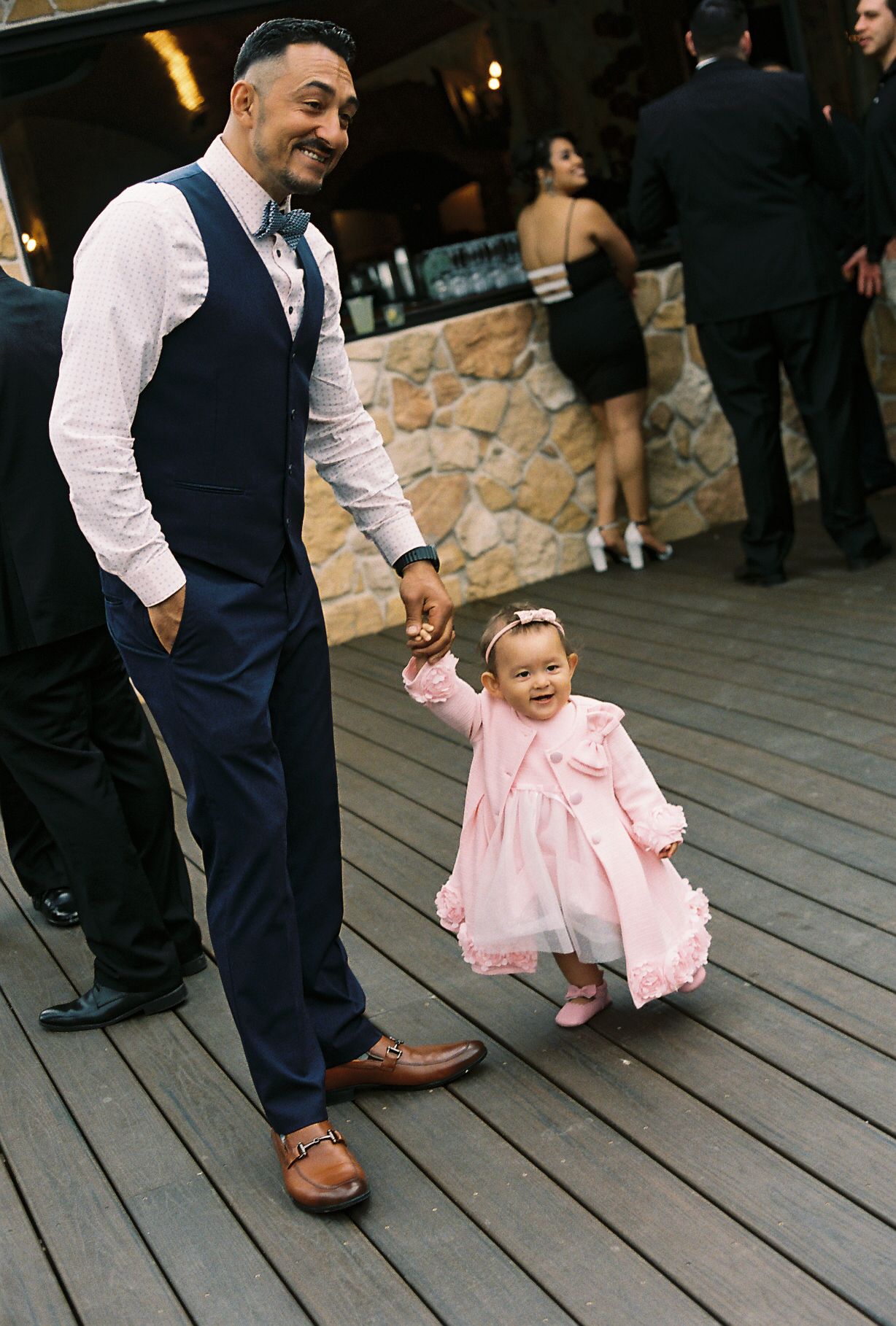 Smiling man in navy vest holds hands with adorable baby girl in pink dress at wedding.