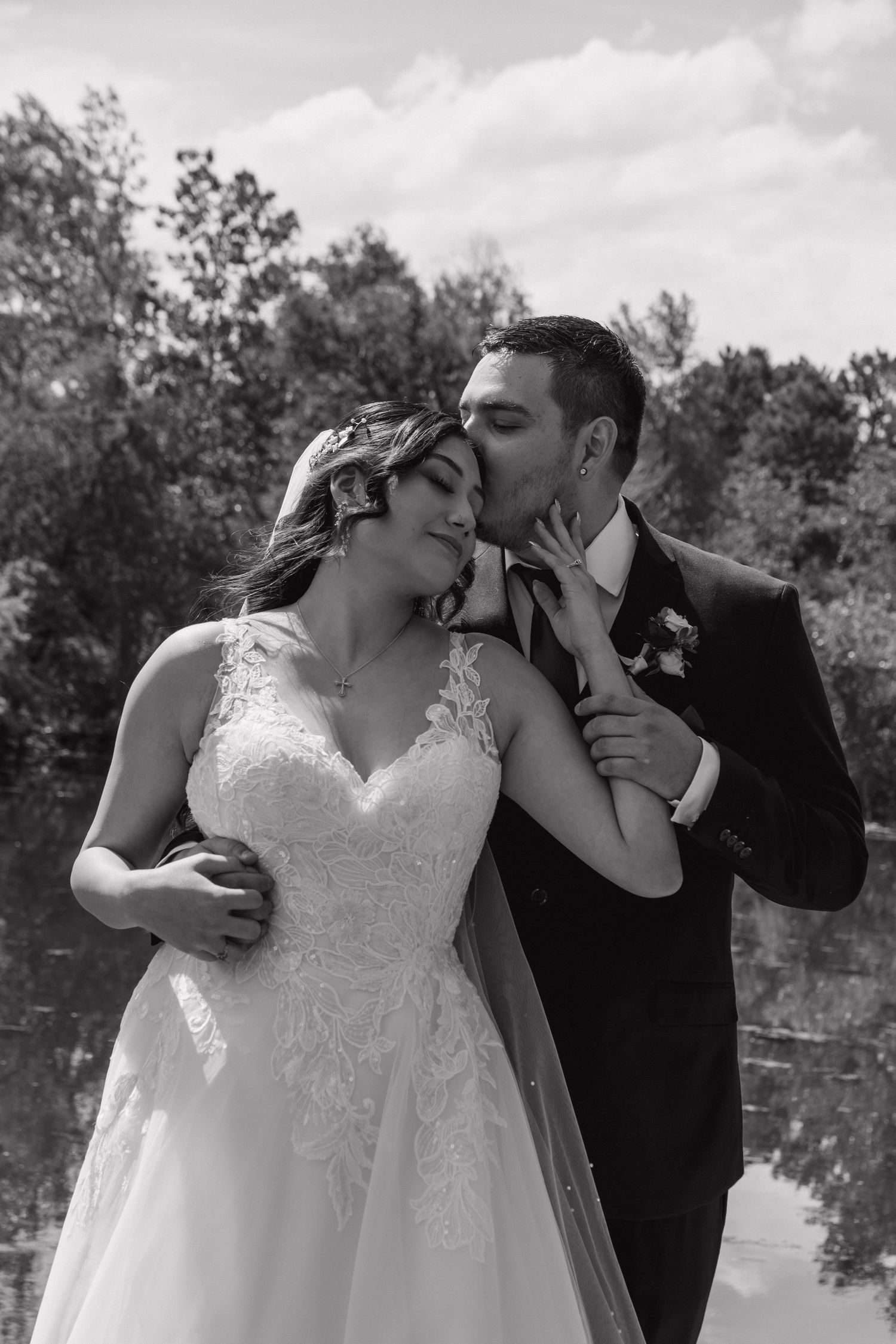Black and white photo of groom kissing bride on cheek outdoors near a reflective pond surrounded by lush trees.