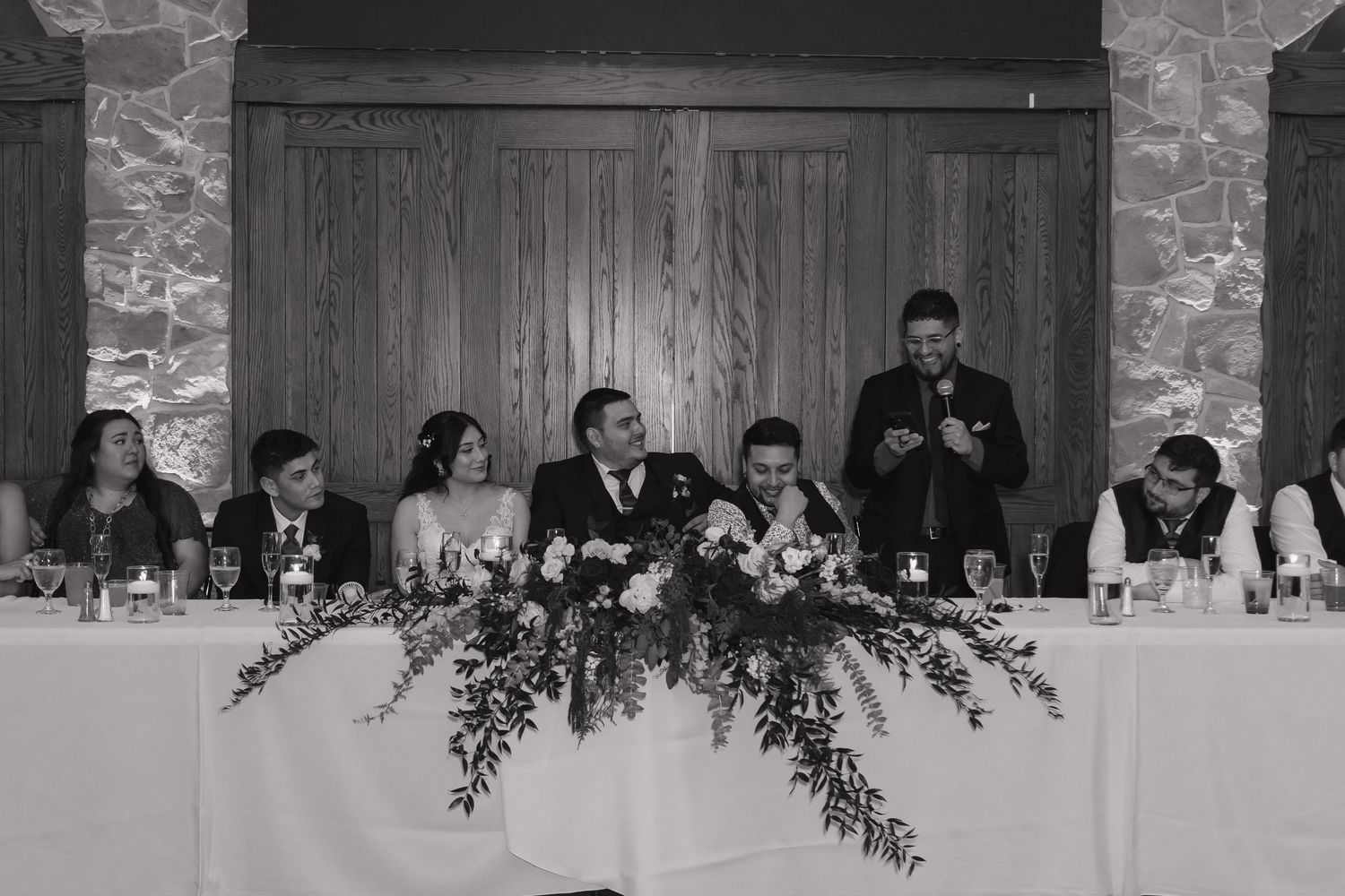 Black and white photo of wedding party seated at the reception head table during speeches.