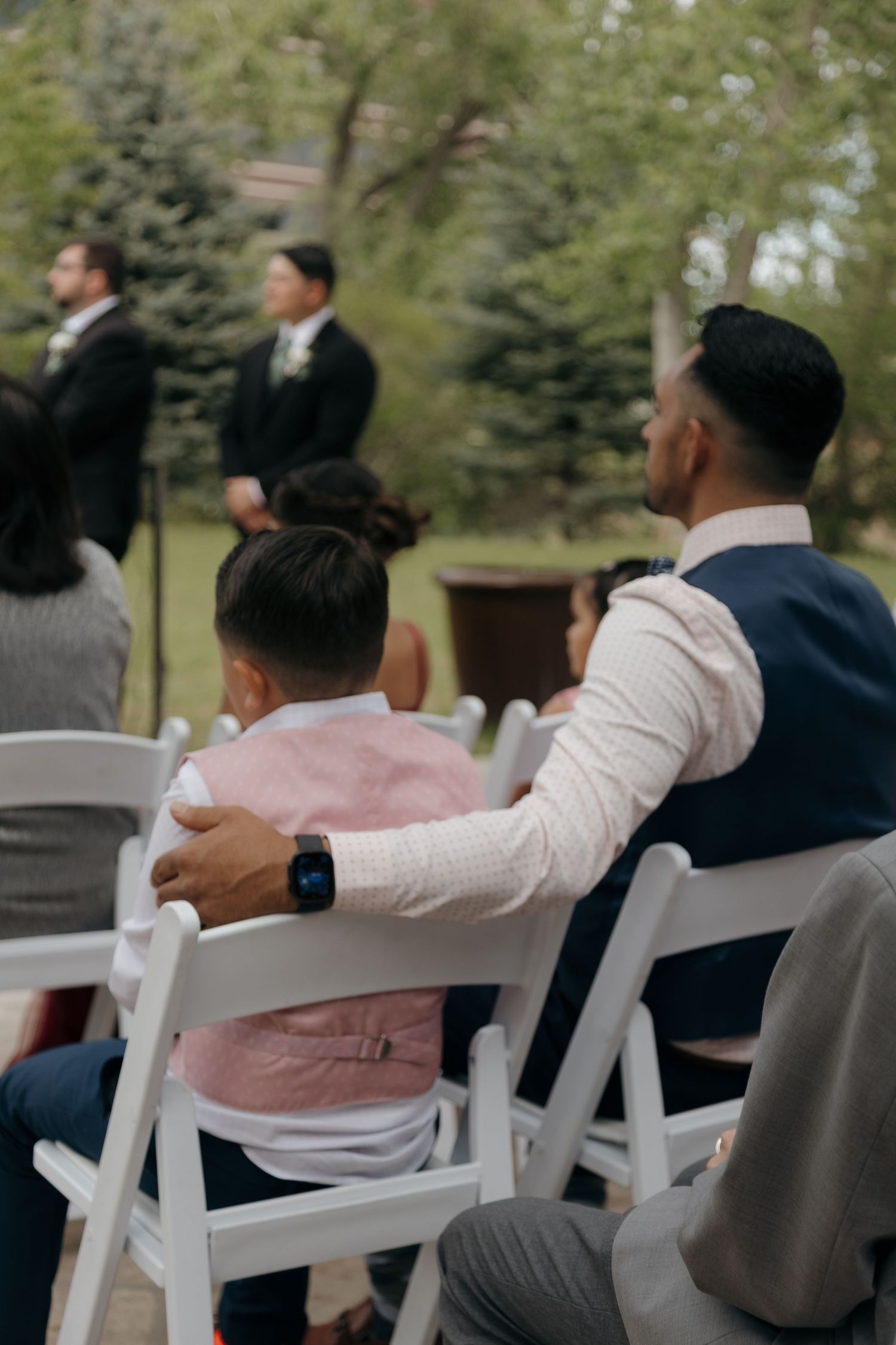 Father placing arm around son while watching outdoor wedding ceremony from reserved seating.
