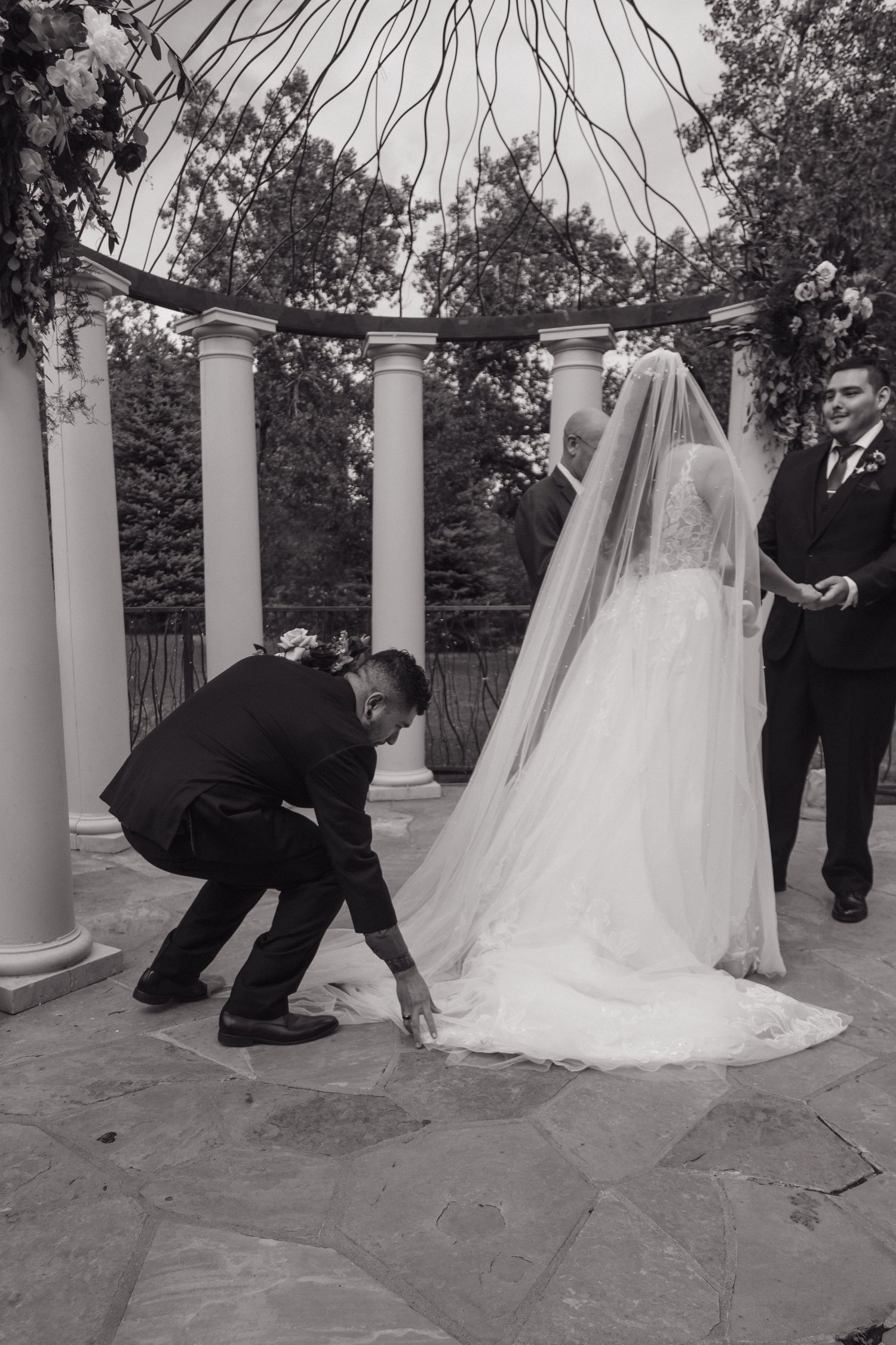 Black and white photo of man adjusting bride's veil train at elegant outdoor wedding ceremony gazebo.