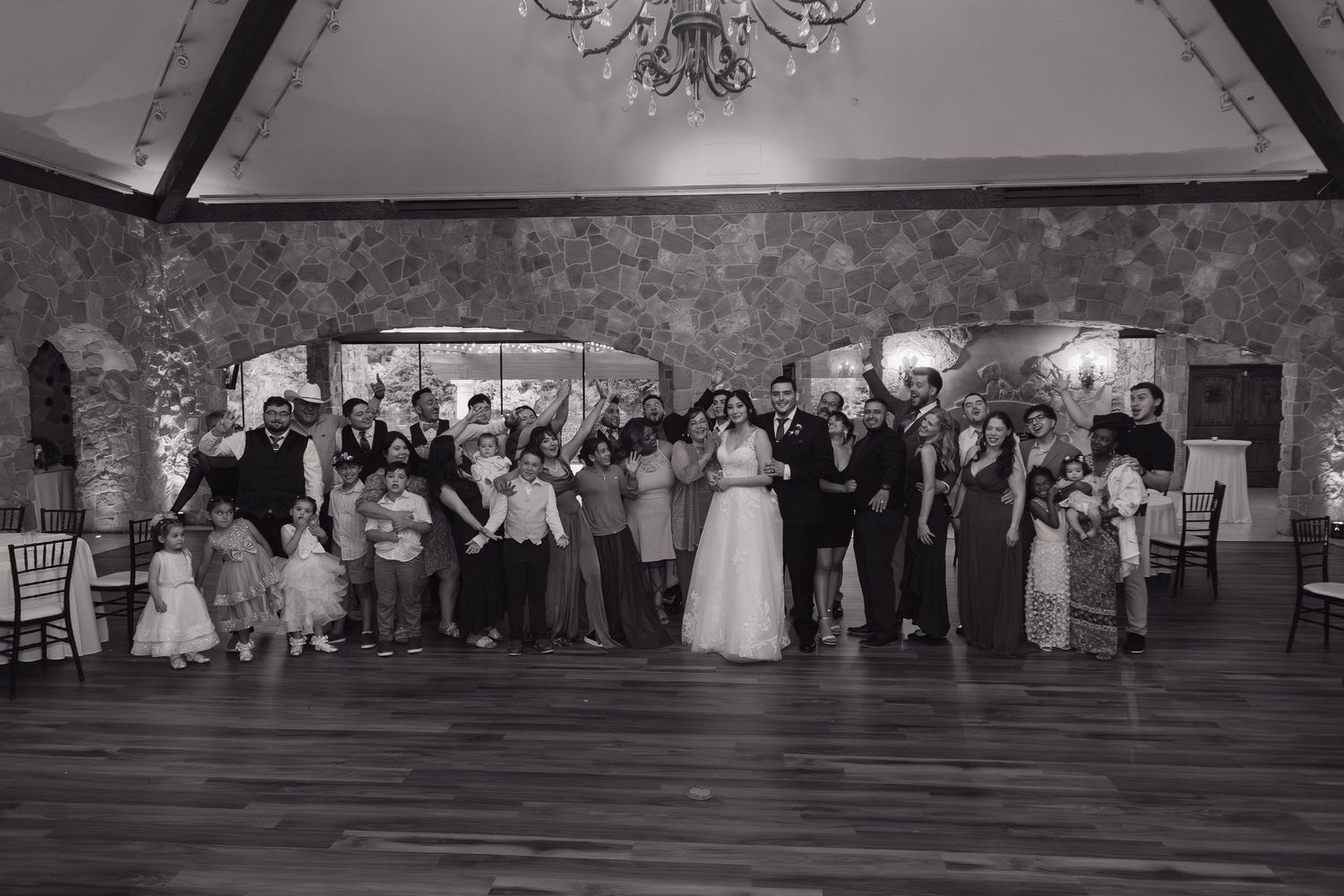 Black and white group photo of entire wedding party on dance floor in elegant venue.
