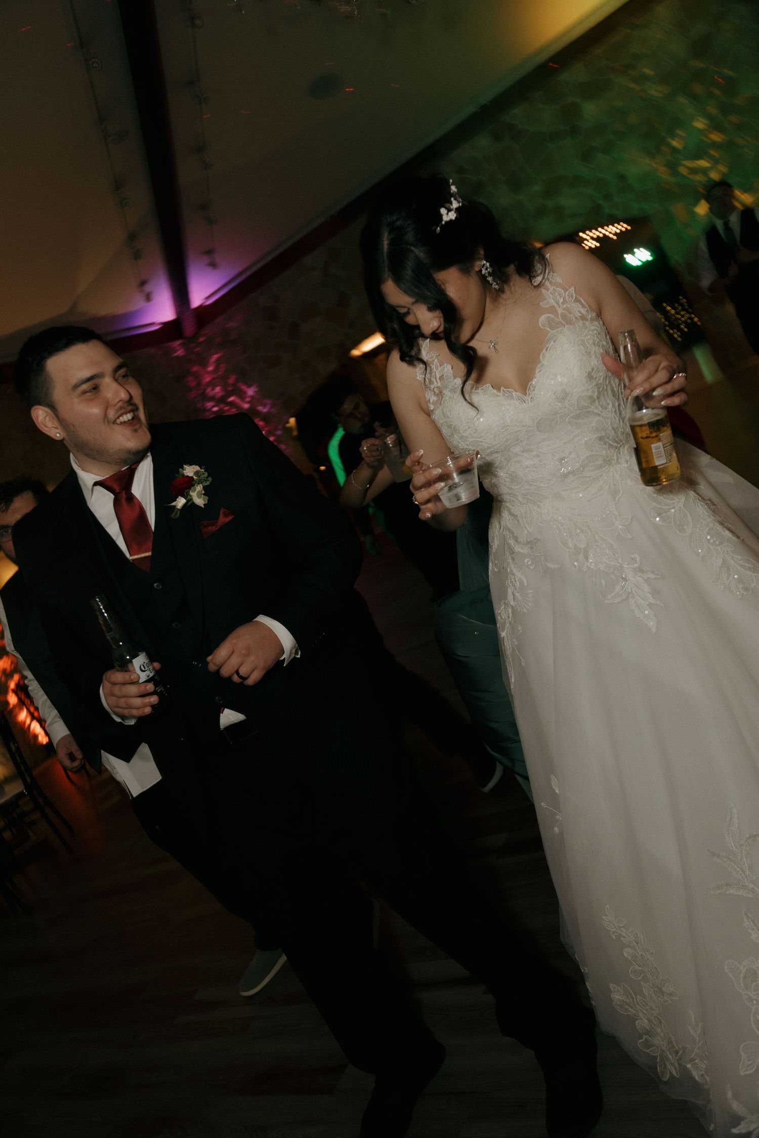 Bride in lace gown and groom in black suit dance joyfully holding drinks at wedding reception.