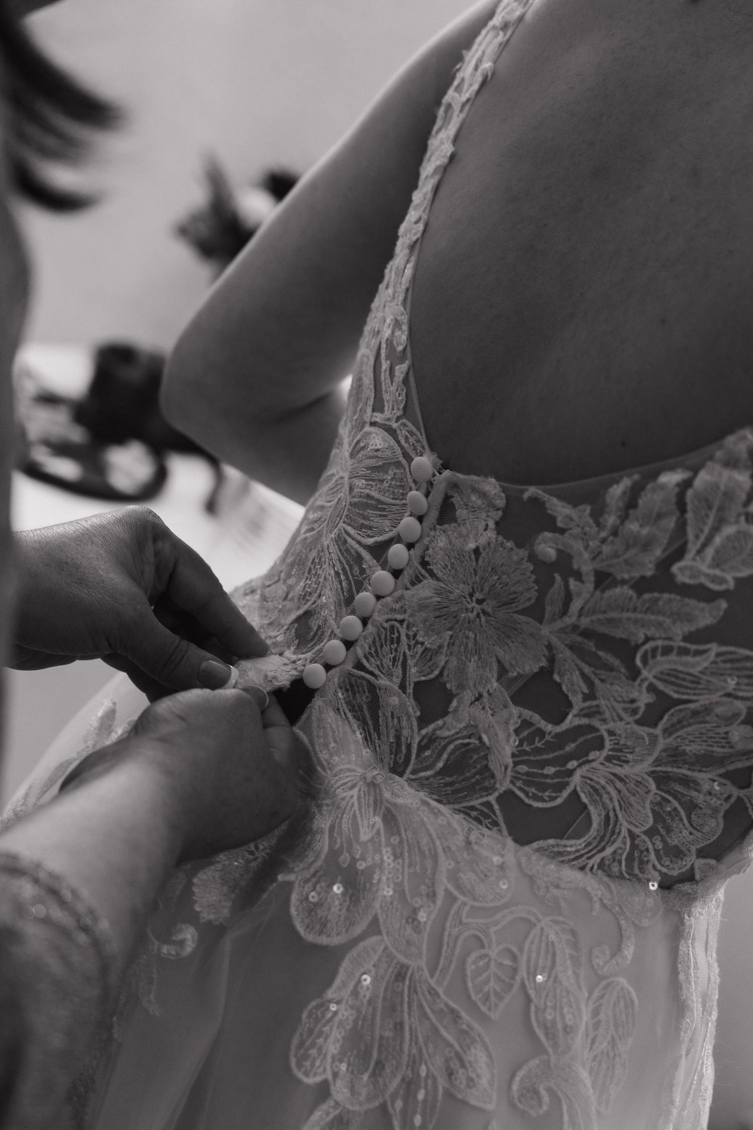 Black and white close-up of hands buttoning intricate lace buttons on the back of a bridal gown.