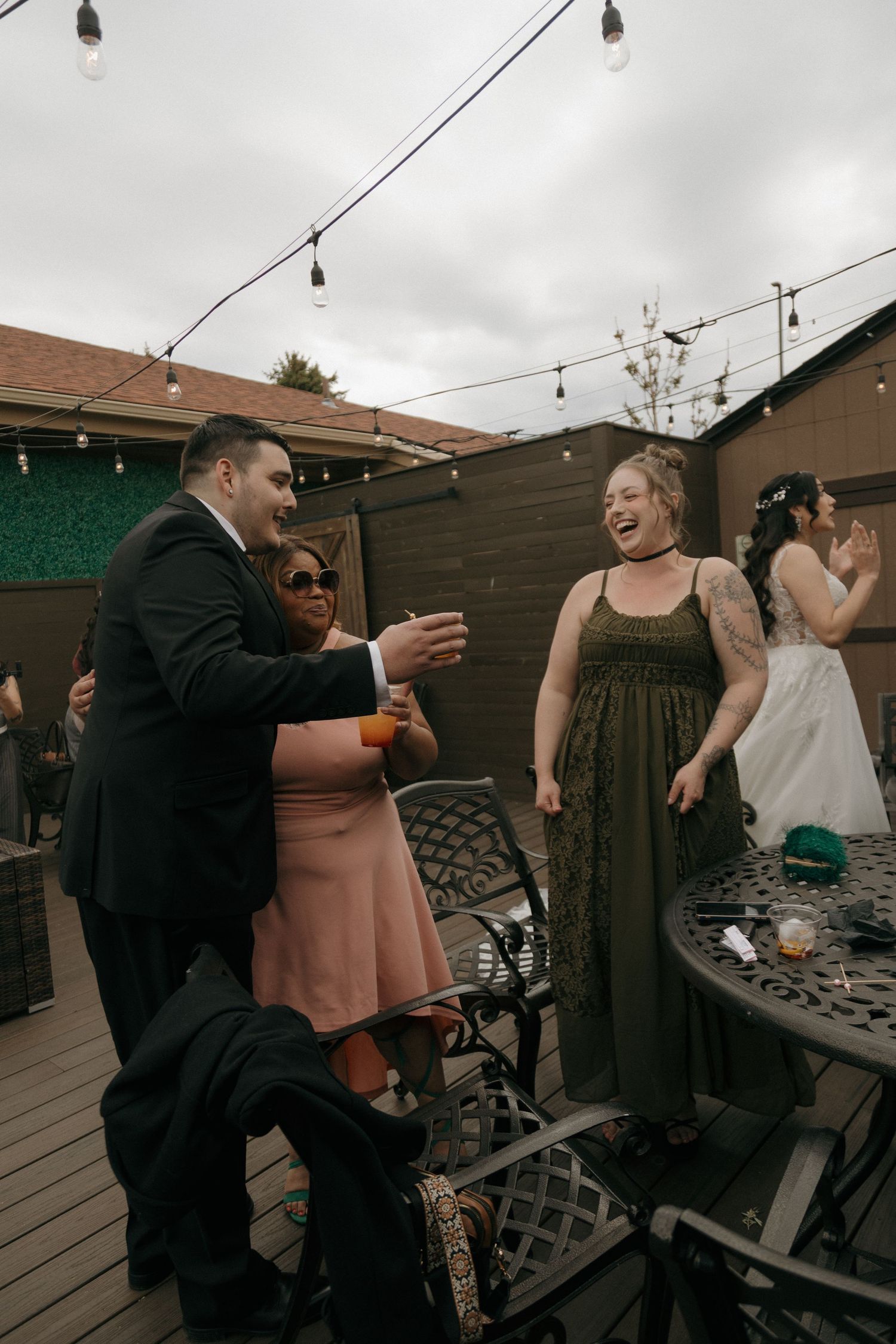 Groom toasts with wedding guests laughing and celebrating at outdoor reception on decorated wooden deck.