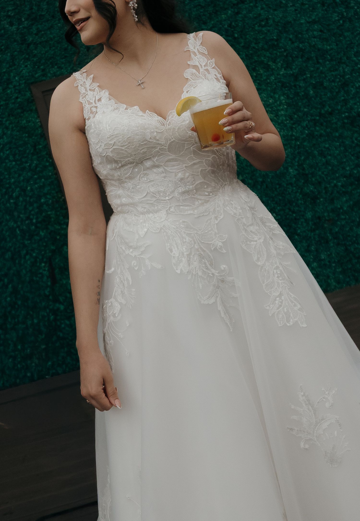 Bride in lace V-neck wedding gown holds a citrus cocktail in front of green hedge wall.