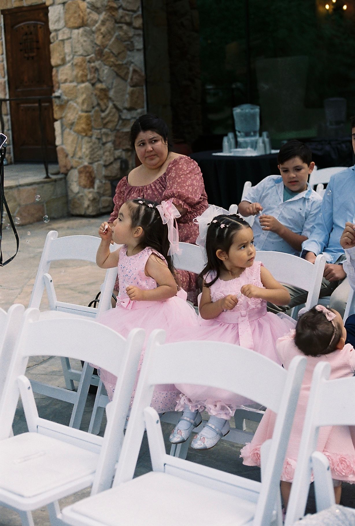 Two little girls in pink dresses sit in white chairs at an outdoor wedding ceremony.