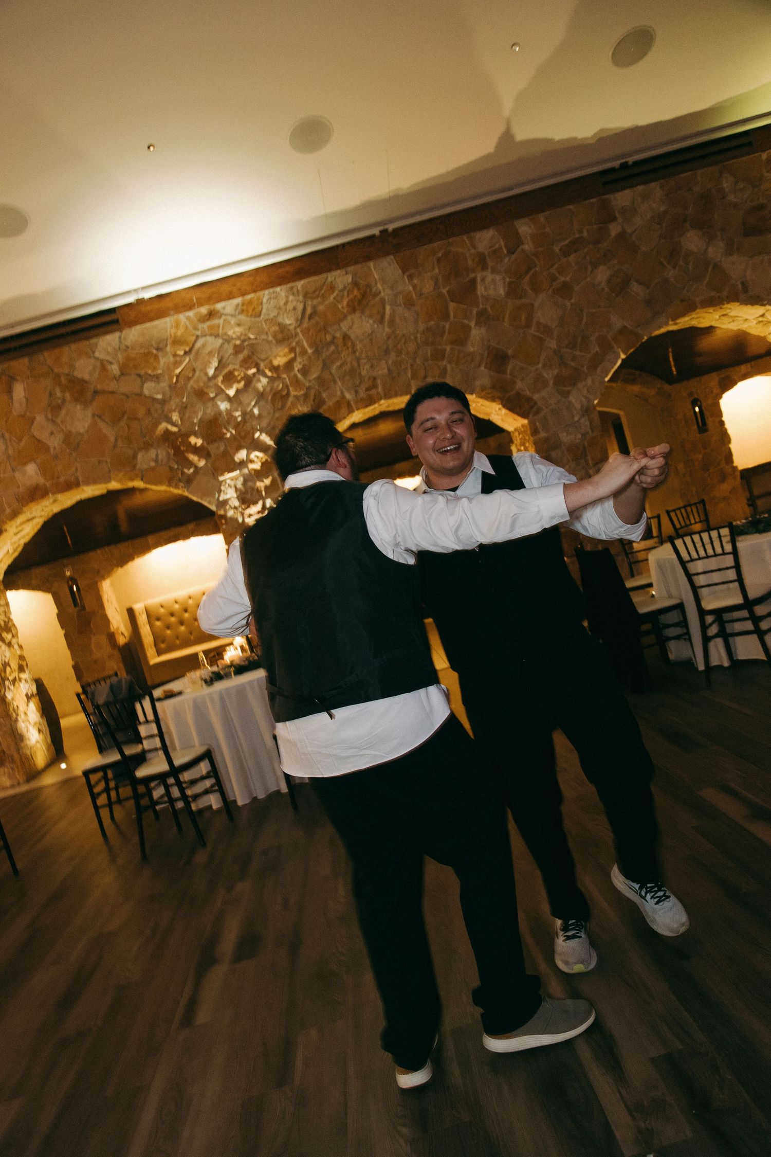 Two groomsmen in black vests dance and celebrate joyfully at wedding reception in stone venue.