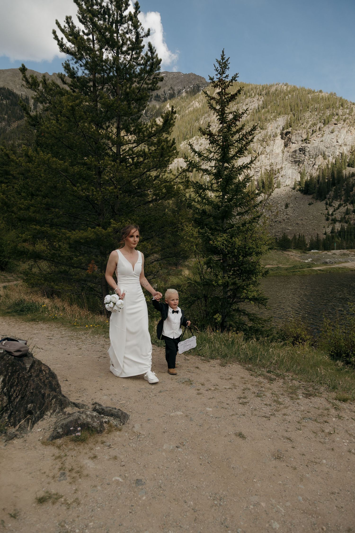 Bride in white gown walks with young child in tuxedo along mountain lake trail.