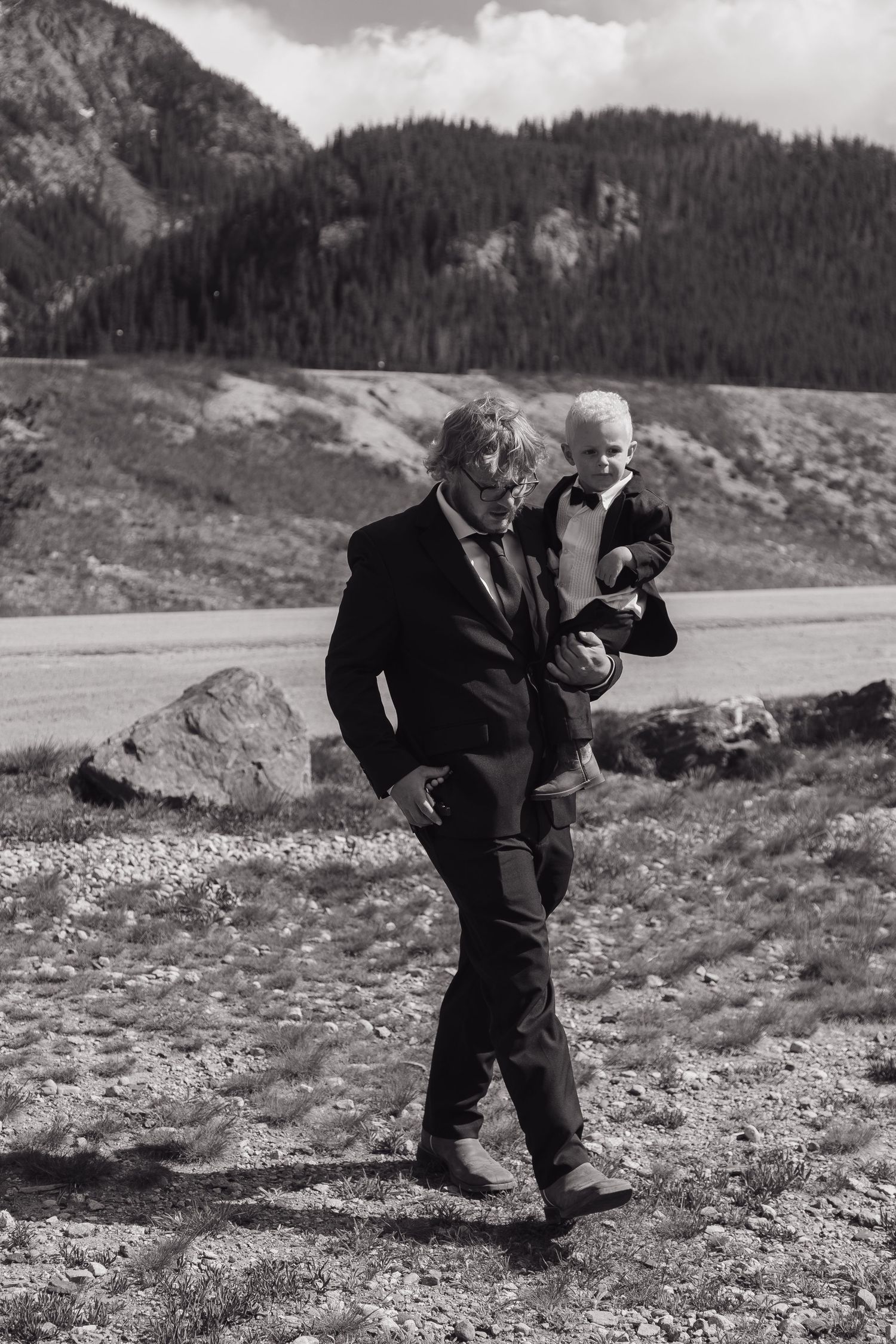 Black and white photo of a man in a suit carrying a young child through a rocky mountain landscape.