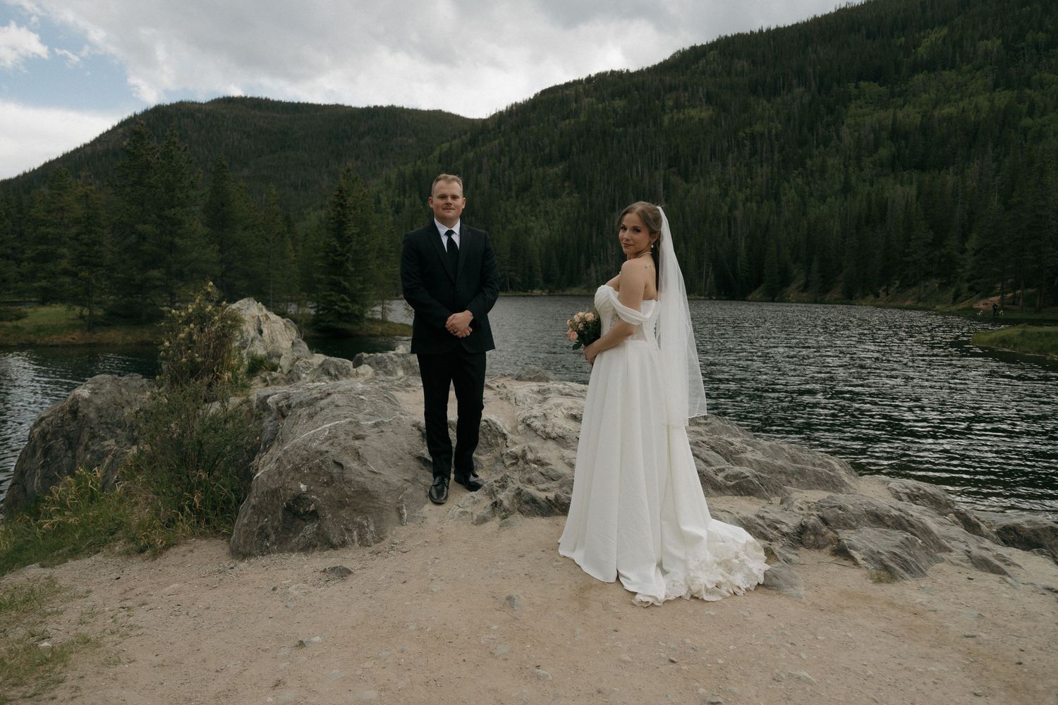 Bride and groom standing on rocky shore beside mountain lake surrounded by pine forest on their wedding day.