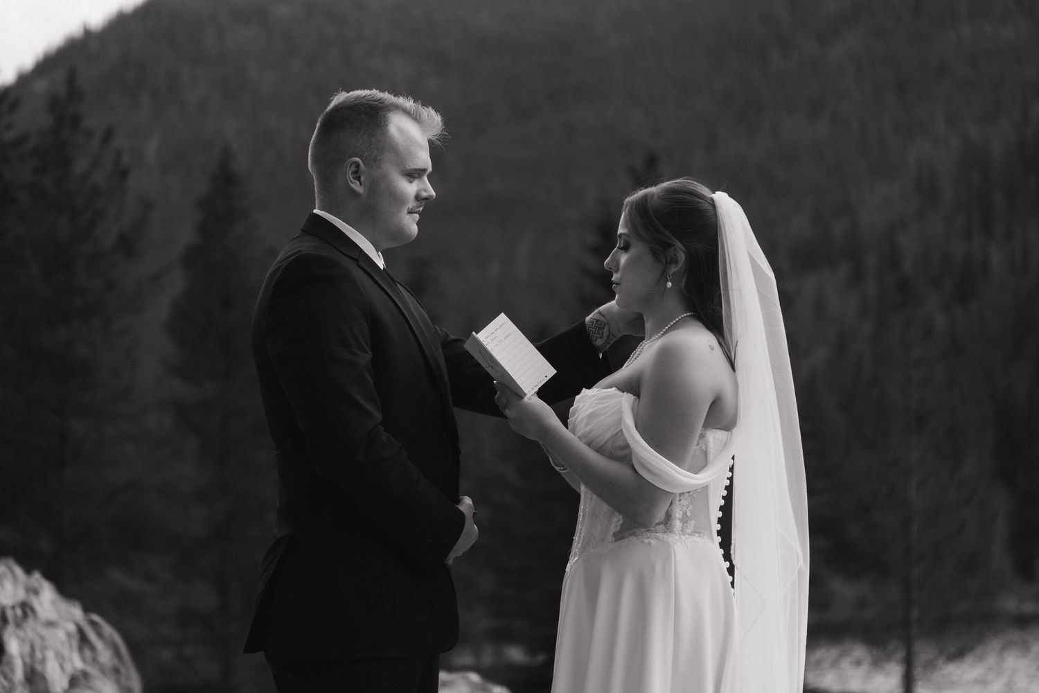 Black and white photo of a bride reading vows to groom outdoors with mountain forest backdrop at wedding ceremony.