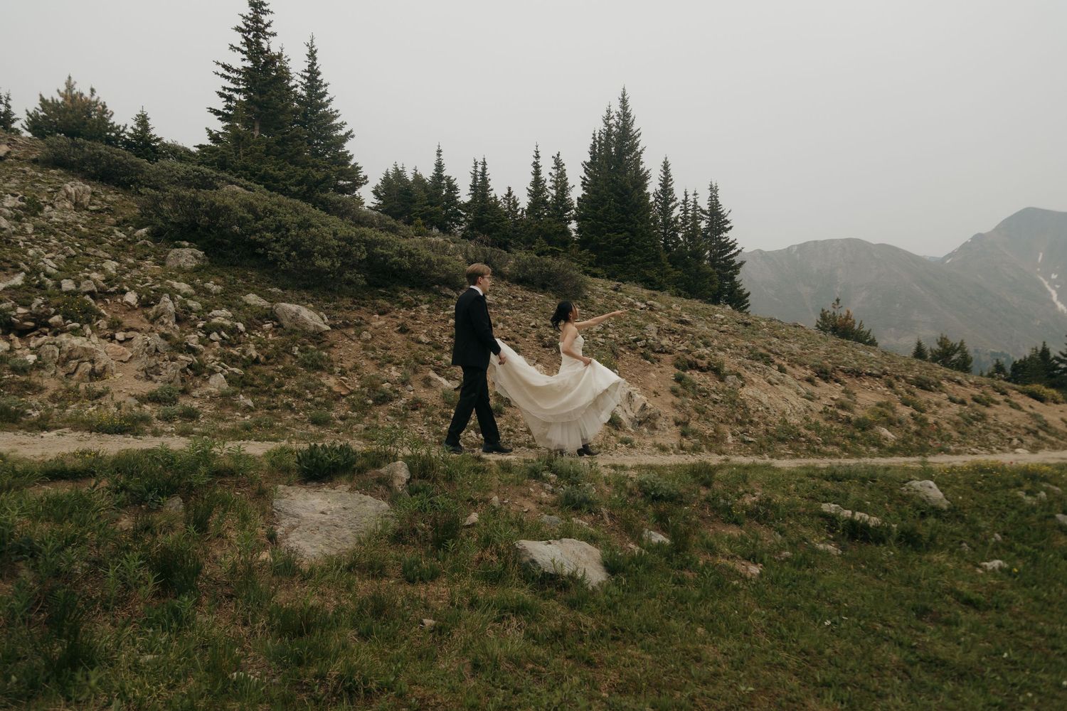 Bride and groom walking a mountain path, her white wedding gown trailing dramatically in the breeze.