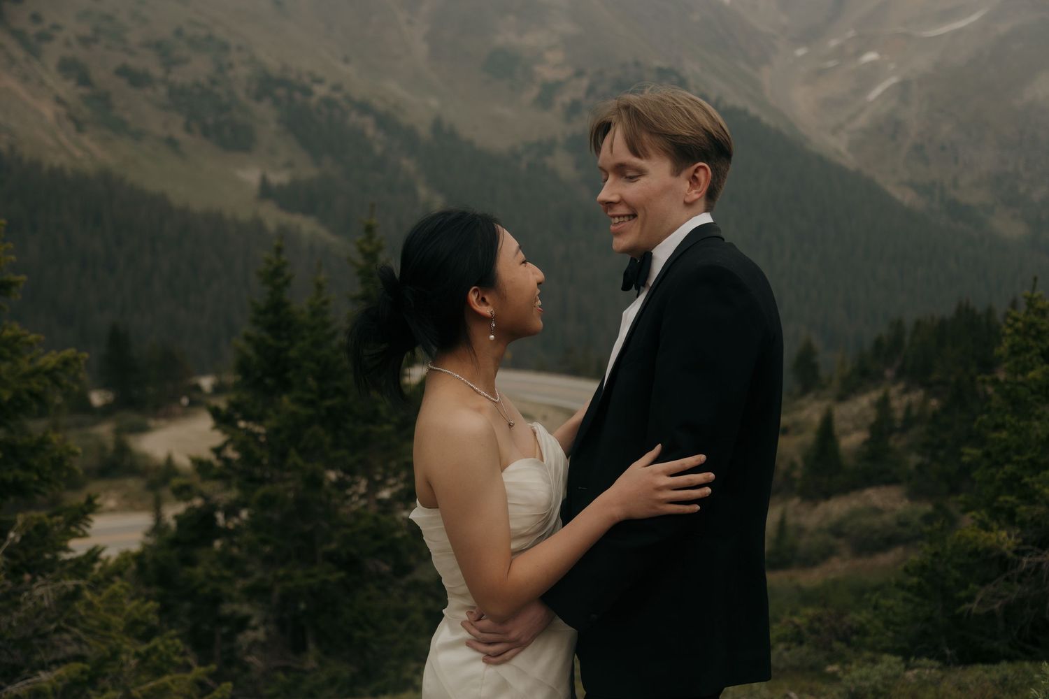 Smiling bride and groom embracing with lush mountain forest backdrop during their outdoor wedding celebration.