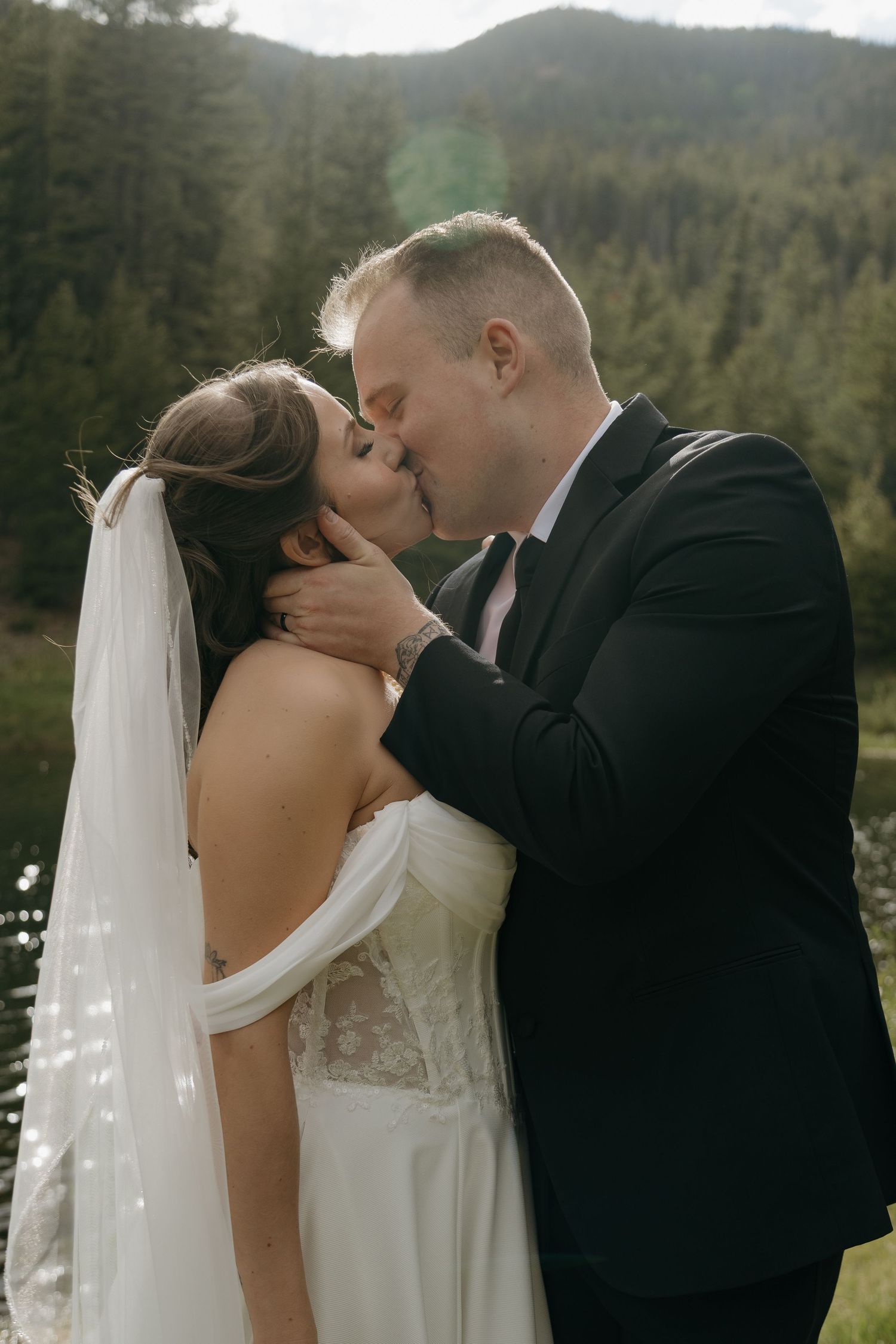 Bride and groom share a romantic kiss with a forested mountain backdrop on their wedding day.