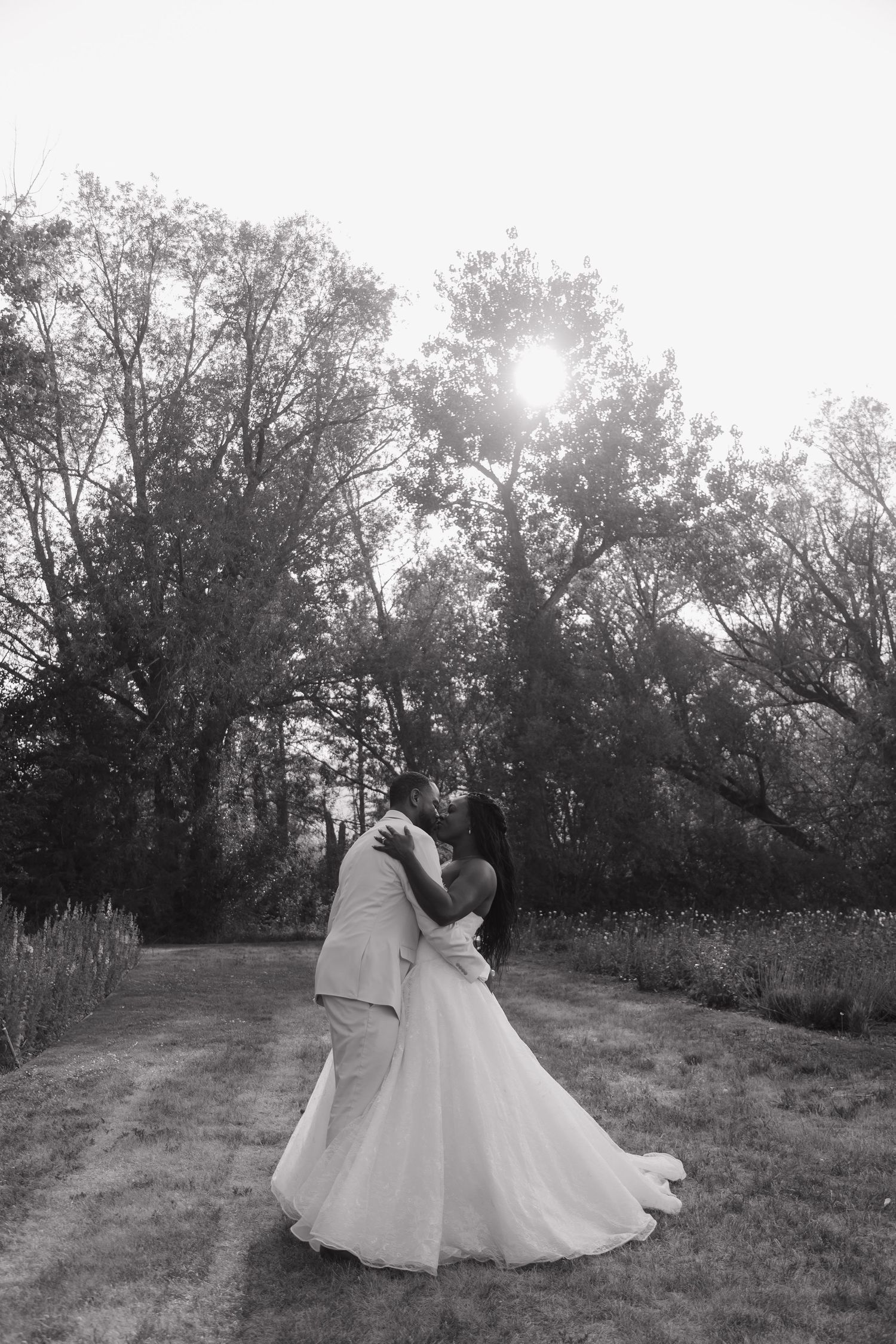 Black and white photo of a bride and groom sharing a romantic kiss outdoors surrounded by tall trees.