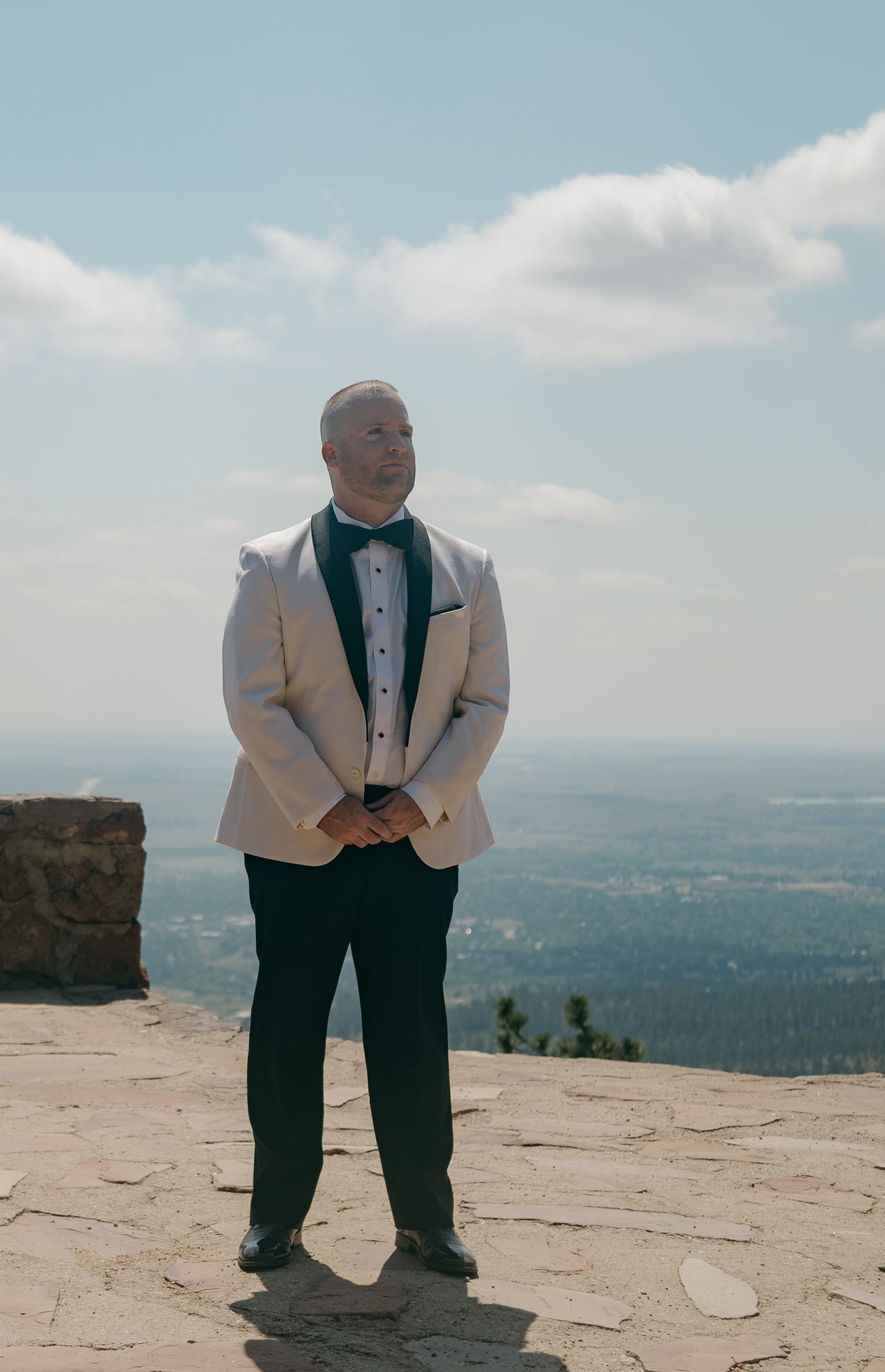 Groom in white tuxedo jacket and bow tie stands on scenic overlook with panoramic view.