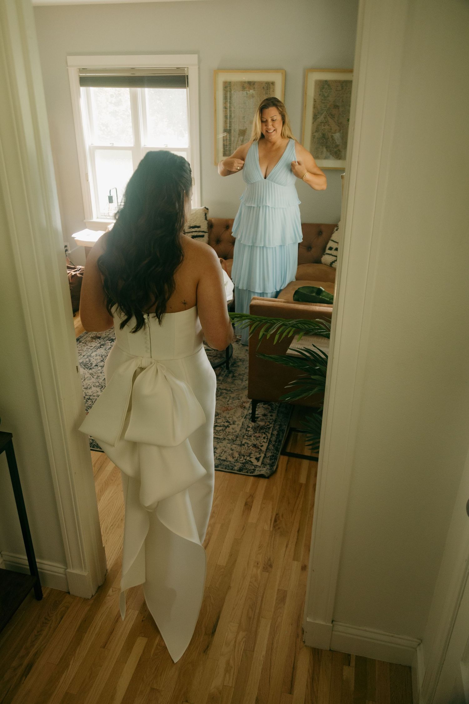 Bride in white gown with bow surprises friend in blue dress during wedding preparation.