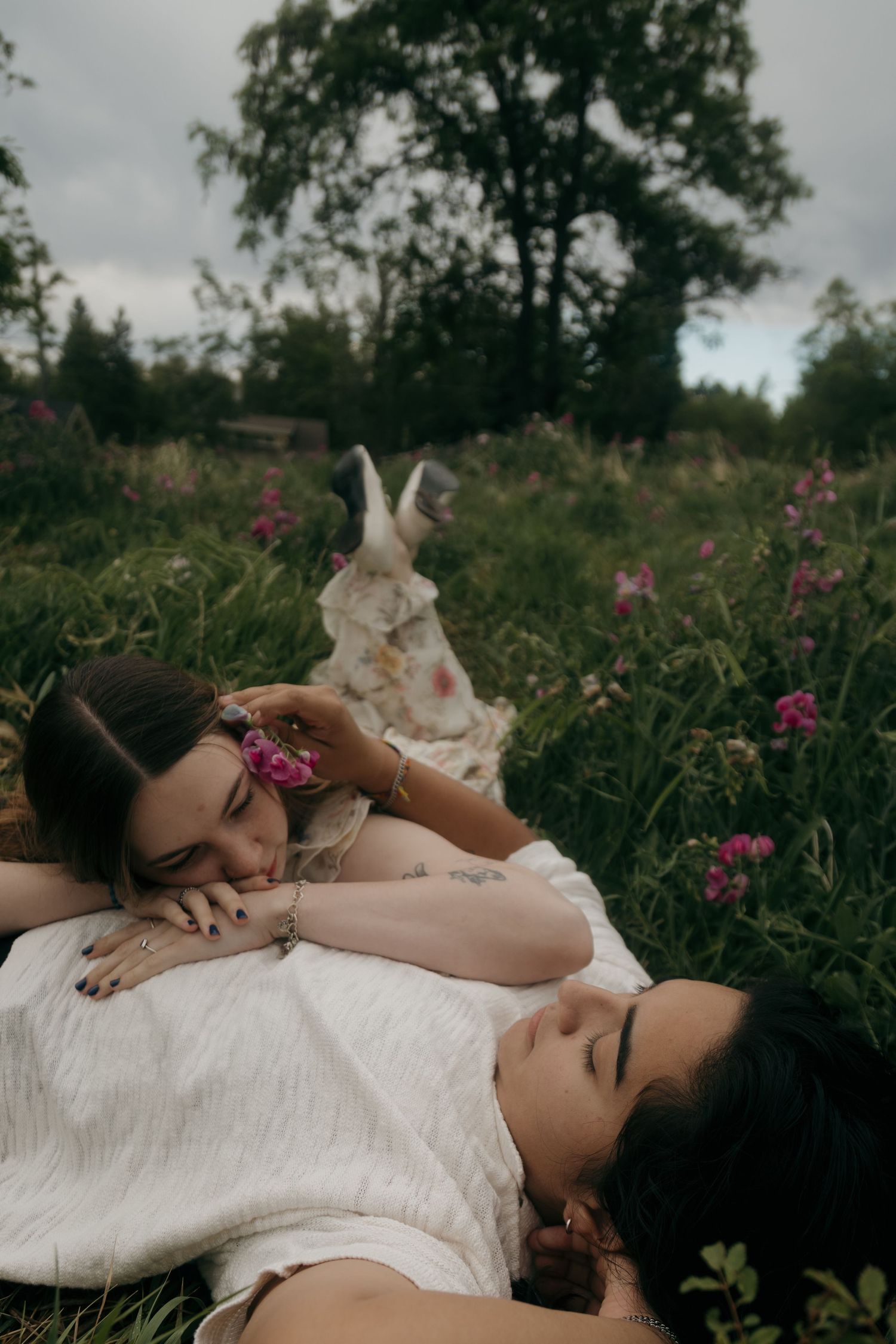 Two women lying in a wildflower meadow, one resting her head on the other's chest tenderly.