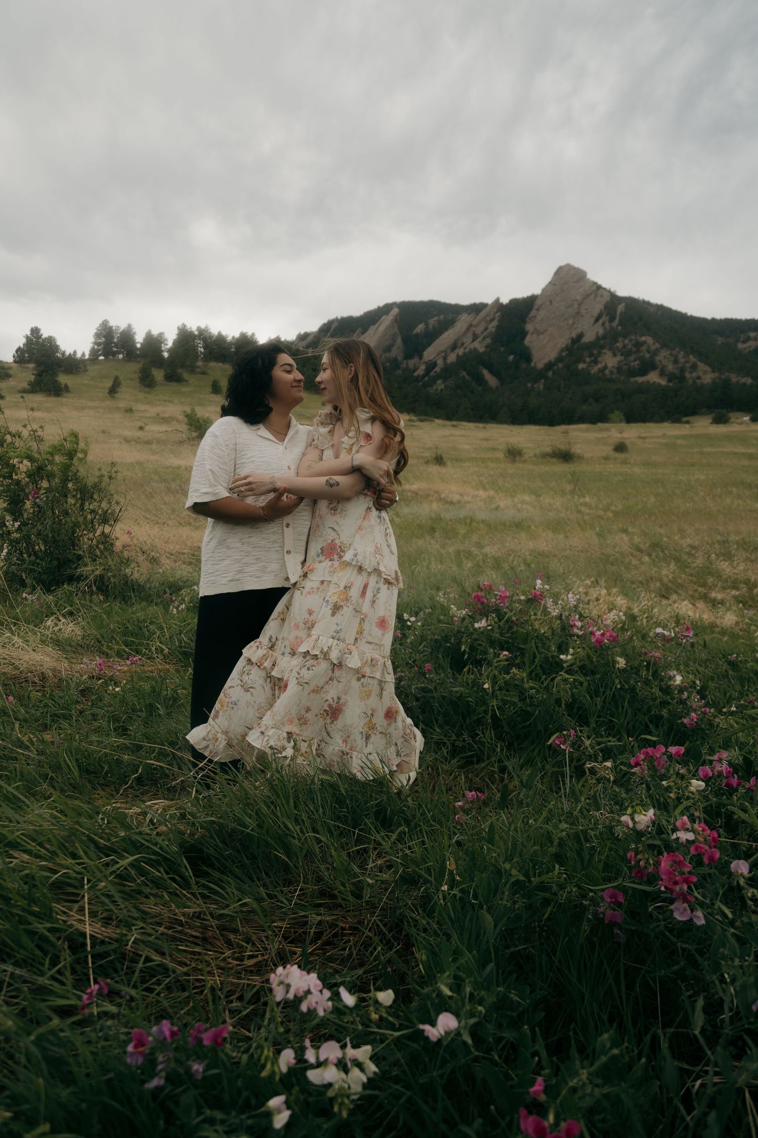 Couple embracing and smiling in a lush green meadow with pink wildflowers and the Flatirons mountains.