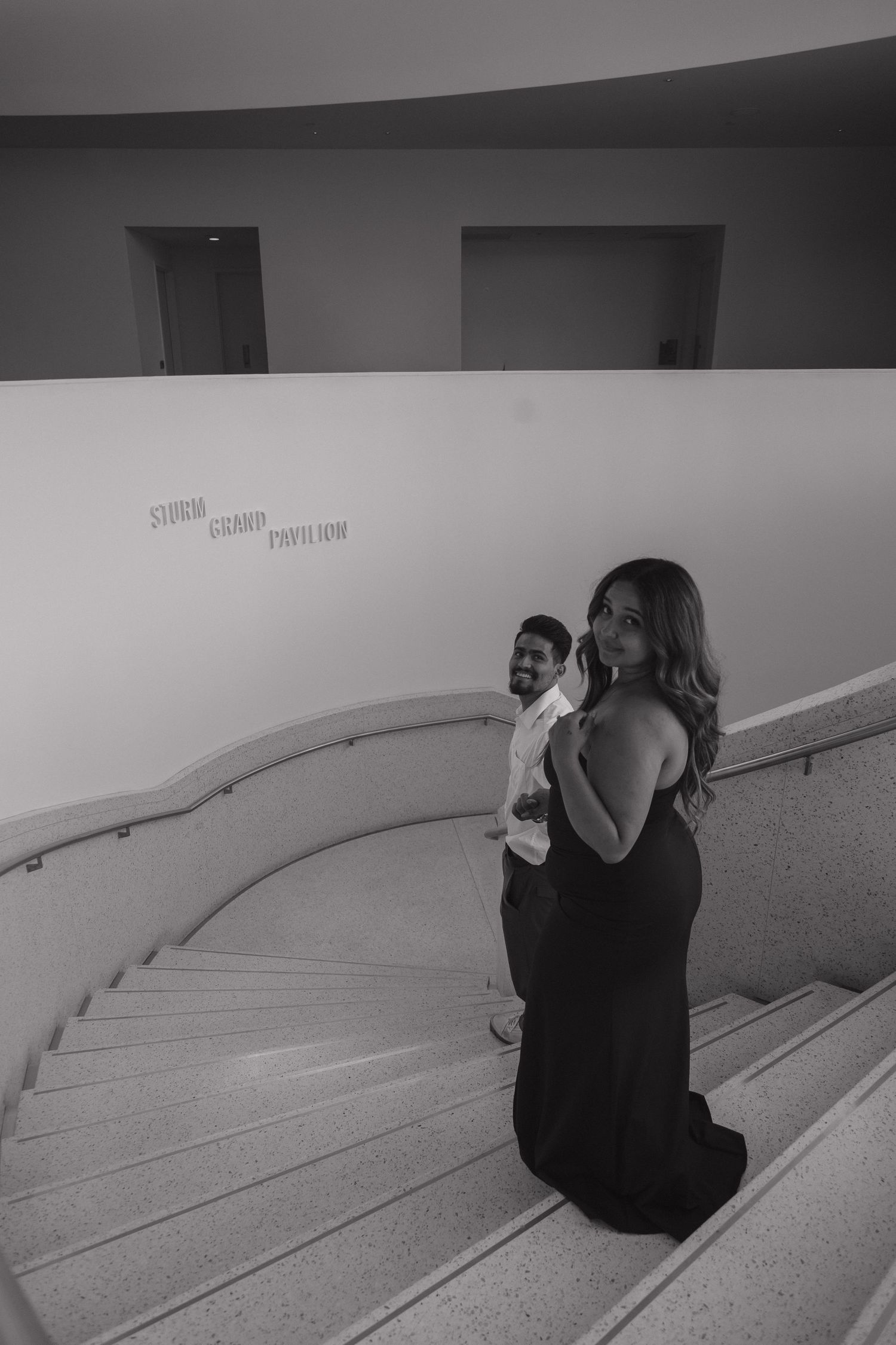 Black and white photo of a smiling couple posing on a curved spiral staircase in a modern building.