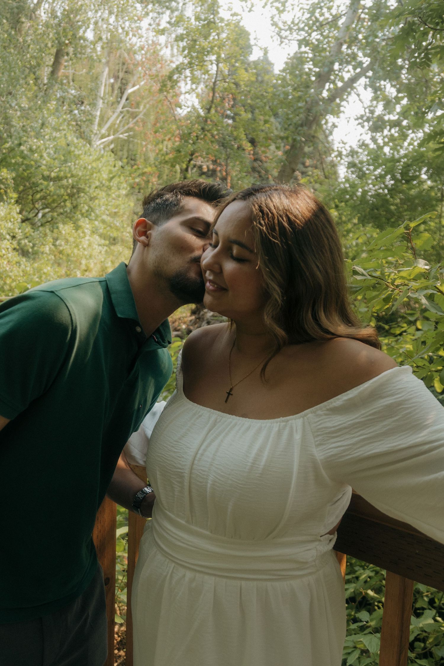 Man in green polo kissing woman in white off-shoulder dress on a wooden deck surrounded by trees.