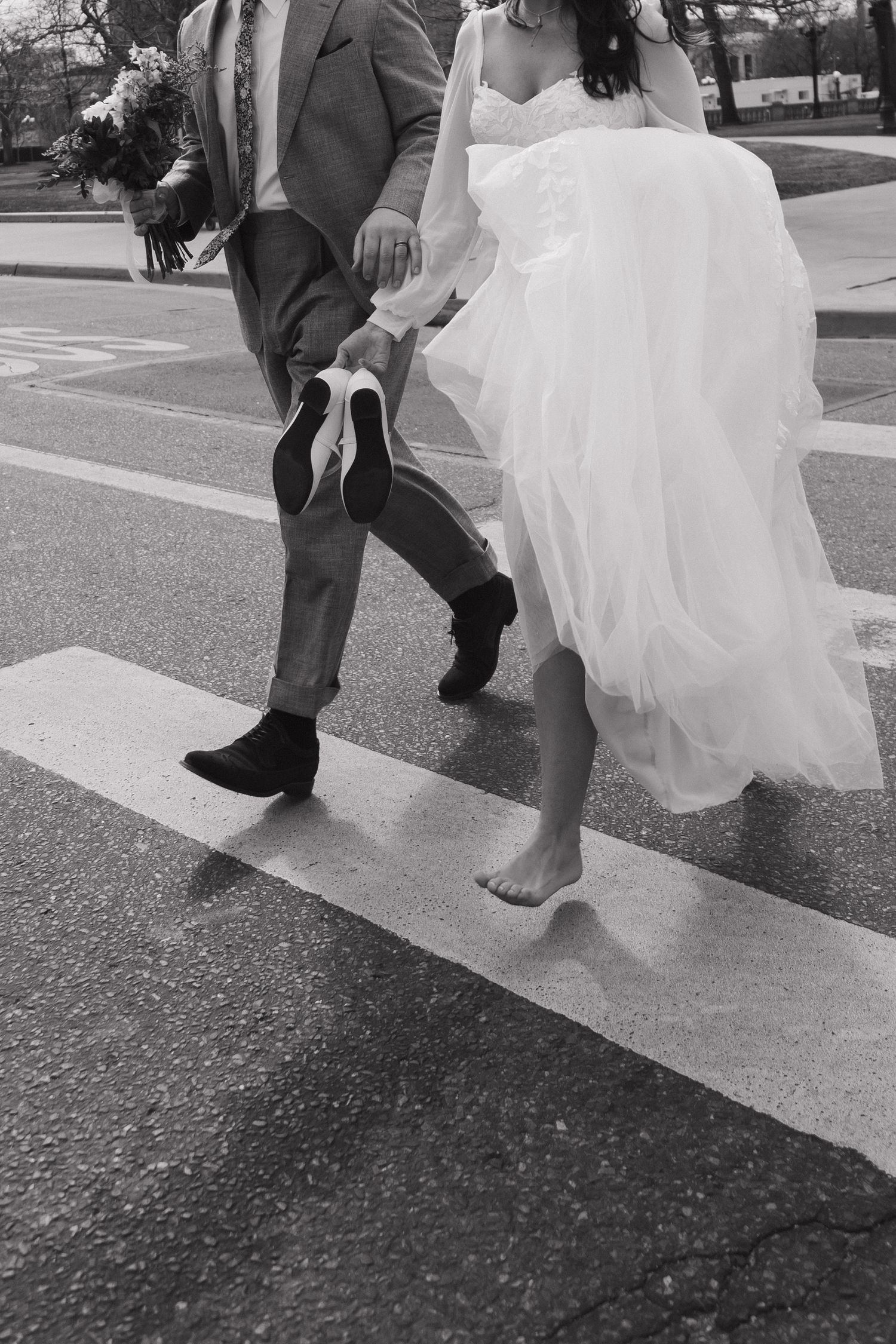 A black and white photo showing the feet of newlyweds crossing a street, with the bride barefoot.