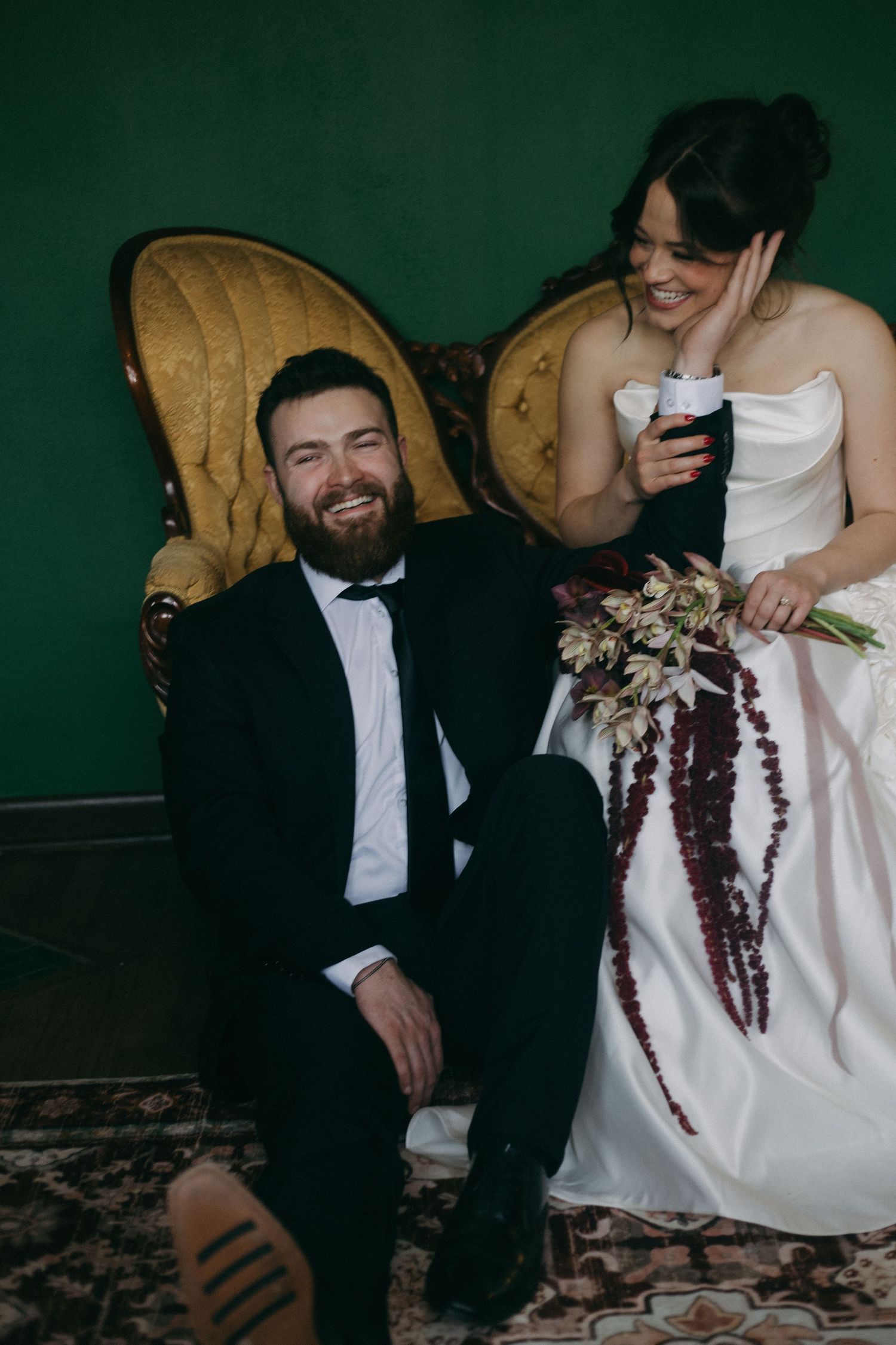 A couple shares a candid moment on a vintage couch with a burgundy floral bouquet.