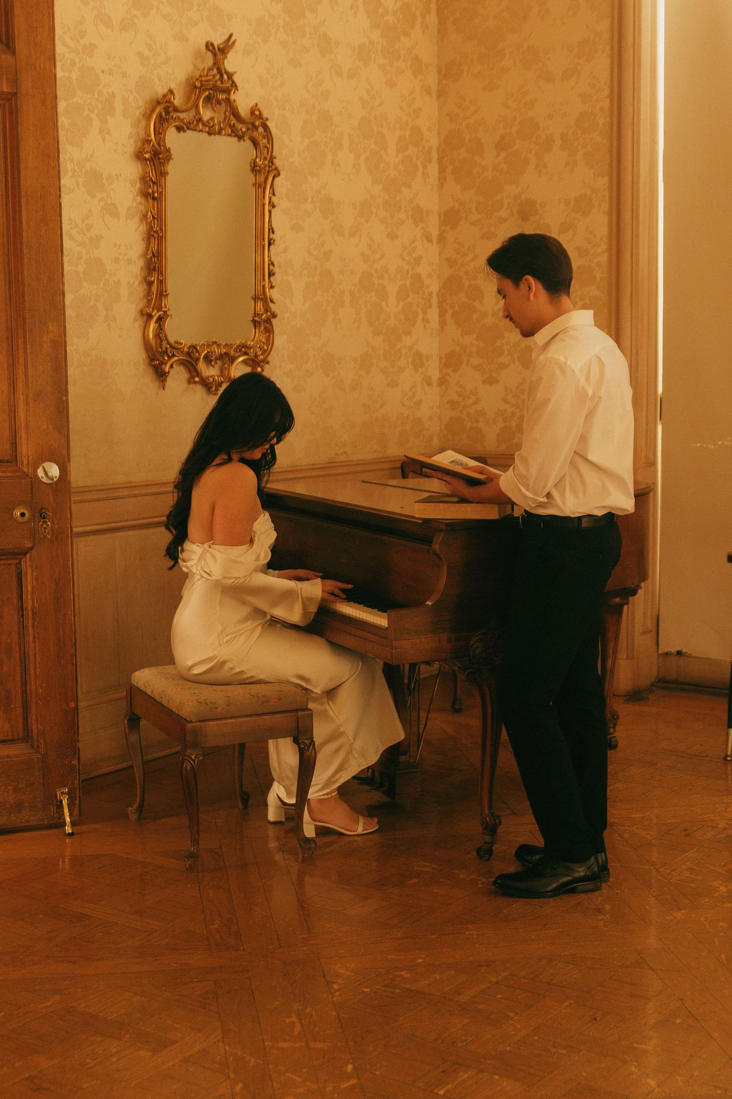 A couple sharing an intimate moment at a piano with an ornate gold mirror on the wall.
