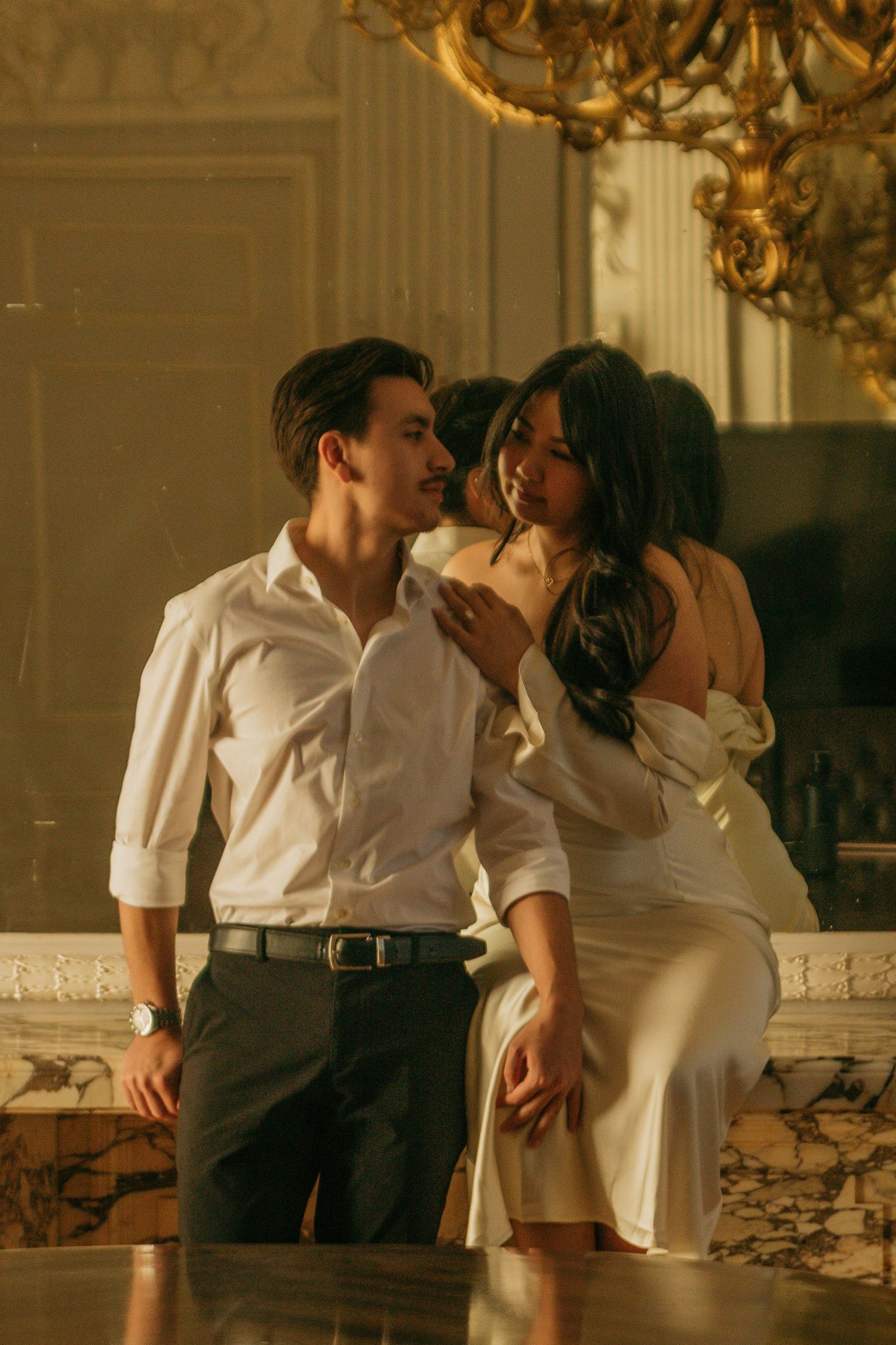A romantic couple in white embraces in front of an ornate chandelier and elegant decor.