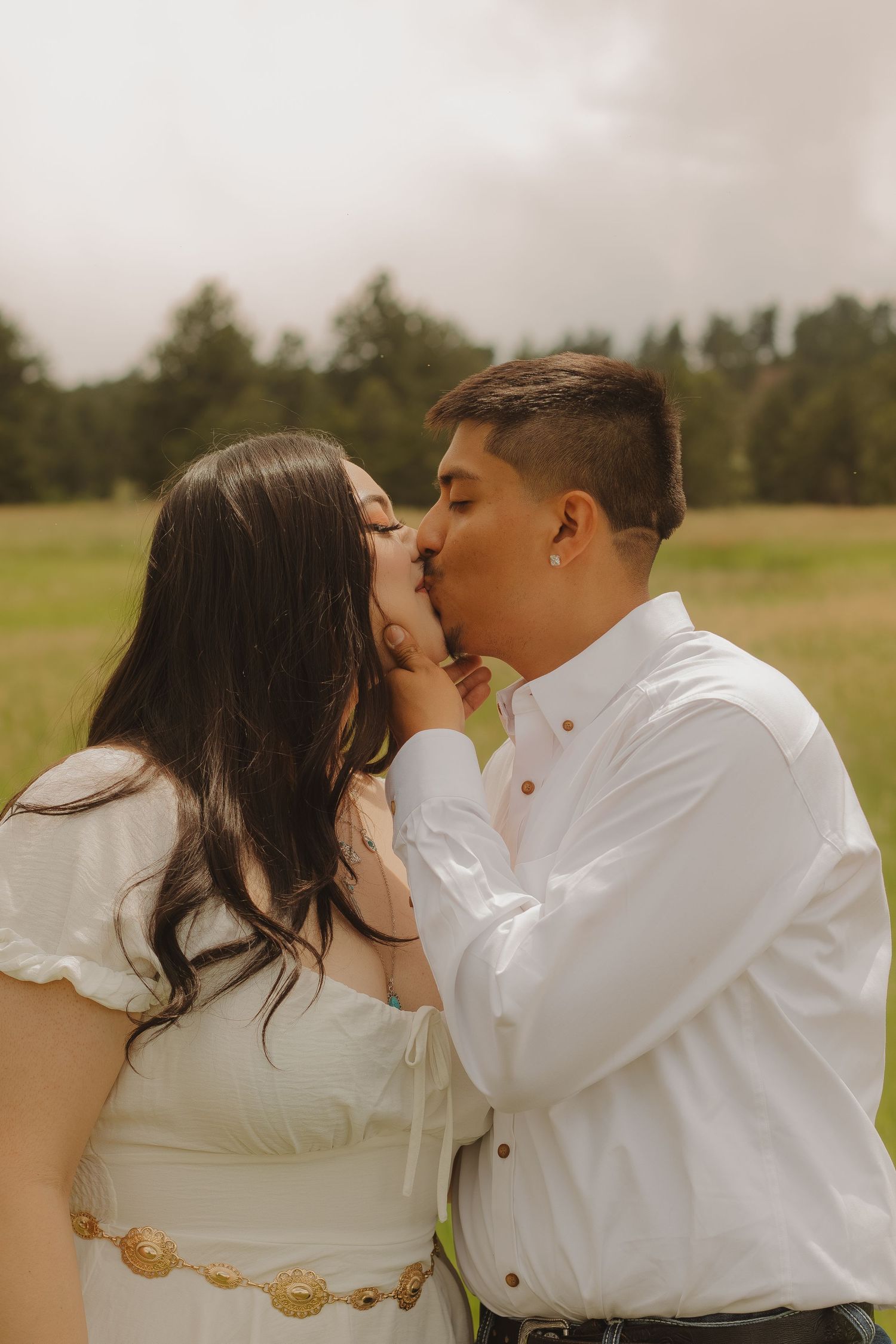 A romantic outdoor photo of a couple sharing a kiss in matching white outfits against a misty natural backdrop.