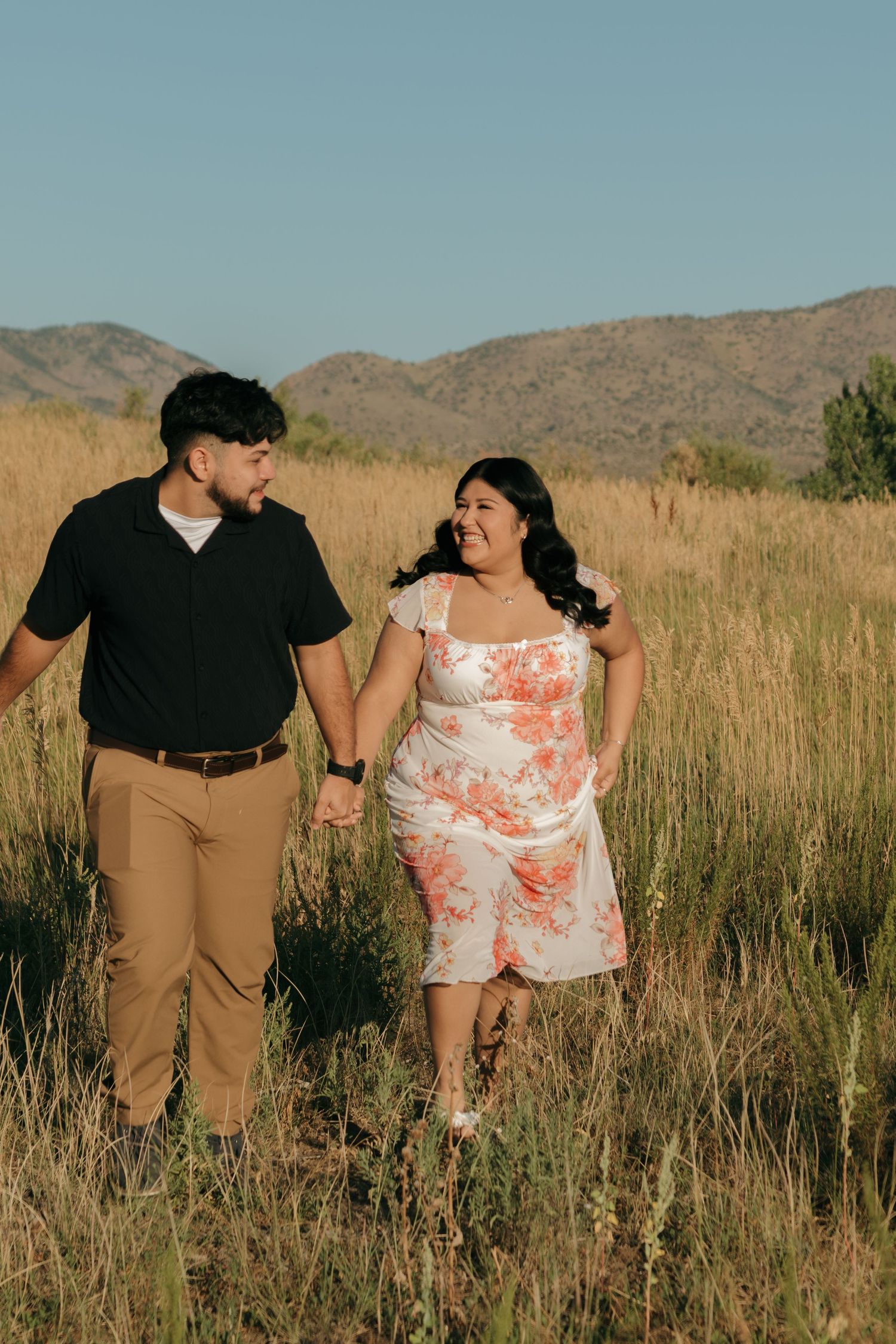 A couple takes a romantic walk through tall grass with mountains in the distance.
