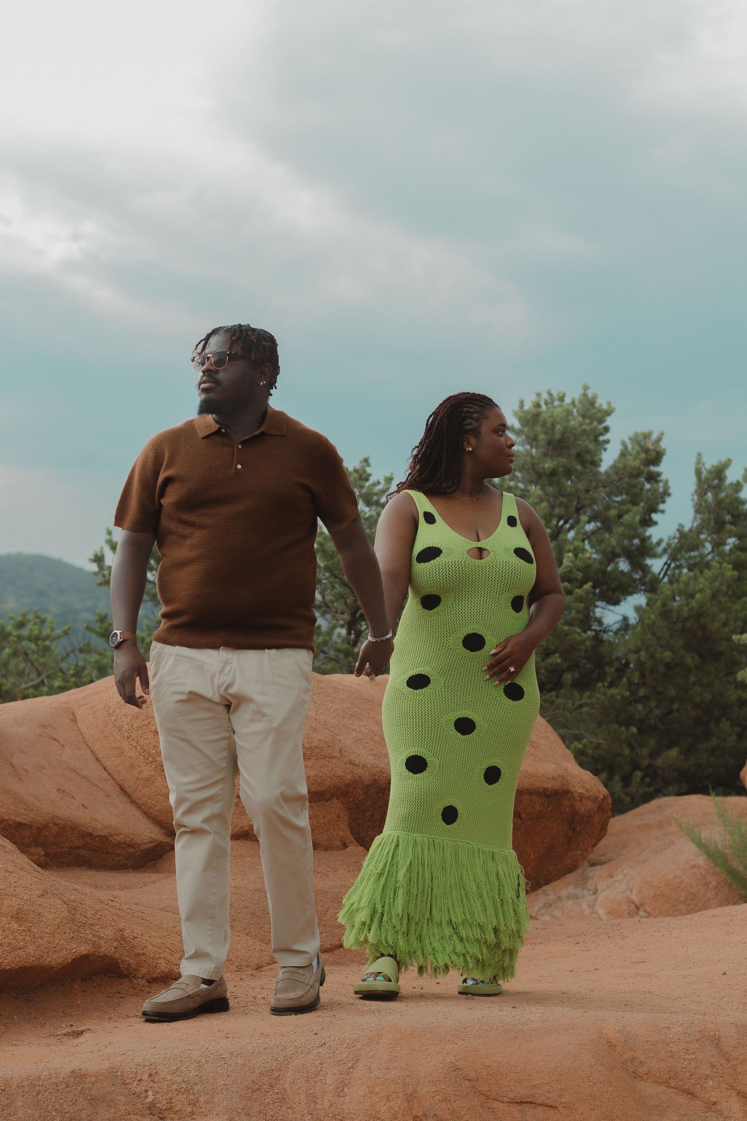 A couple poses together on red rocks against a mountain backdrop, wearing a brown polo and polka dot green dress with fringe.