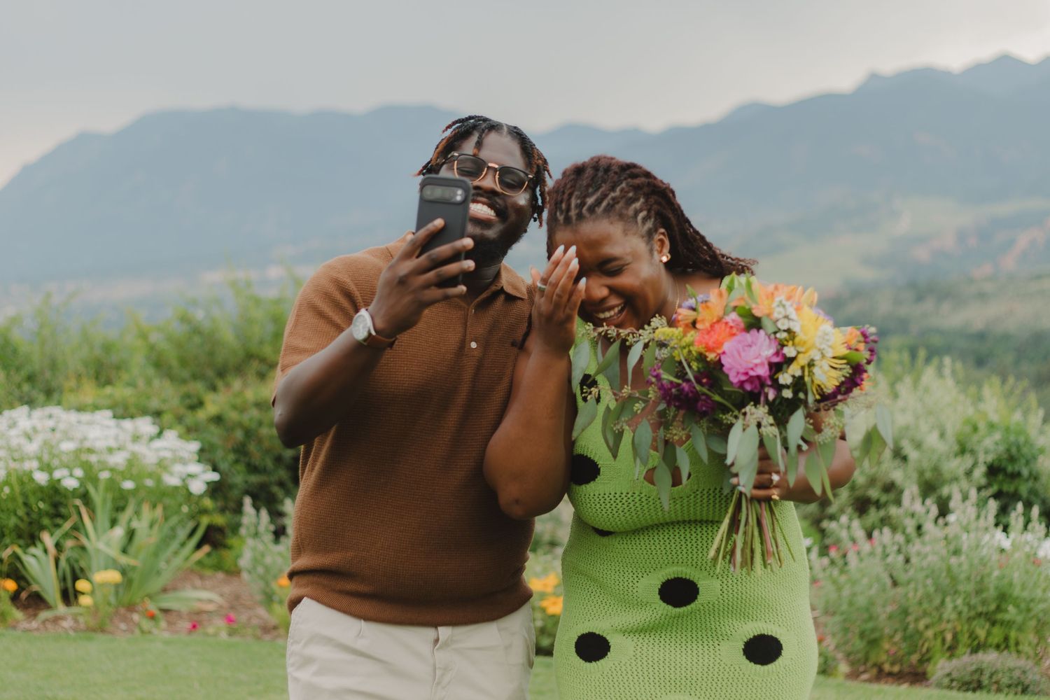 Two friends share a joyful moment outdoors with mountains in background and colorful bouquet of flowers.