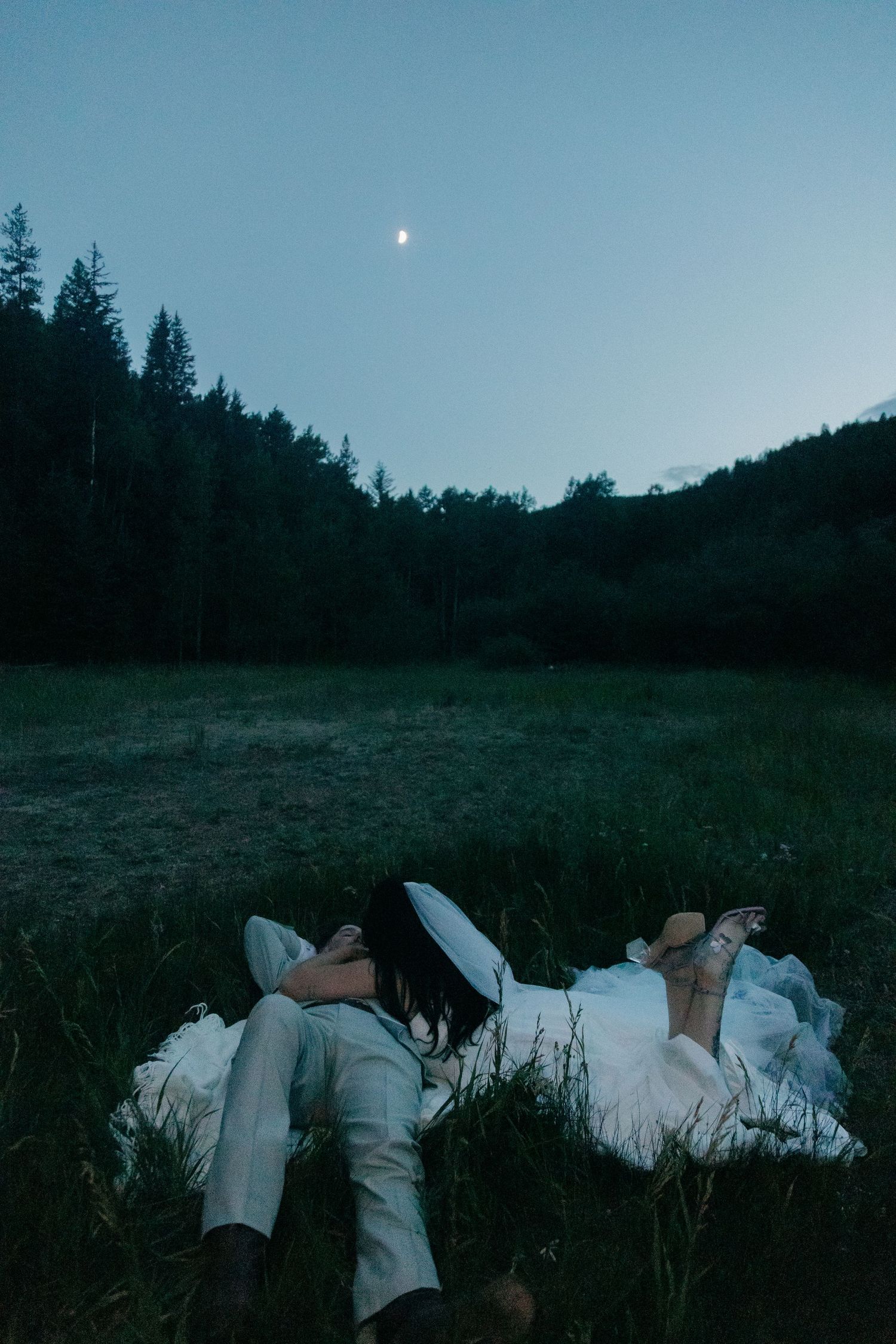 Newlywed couple lying in grass meadow gazing at moon in twilight sky surrounded by forest trees.