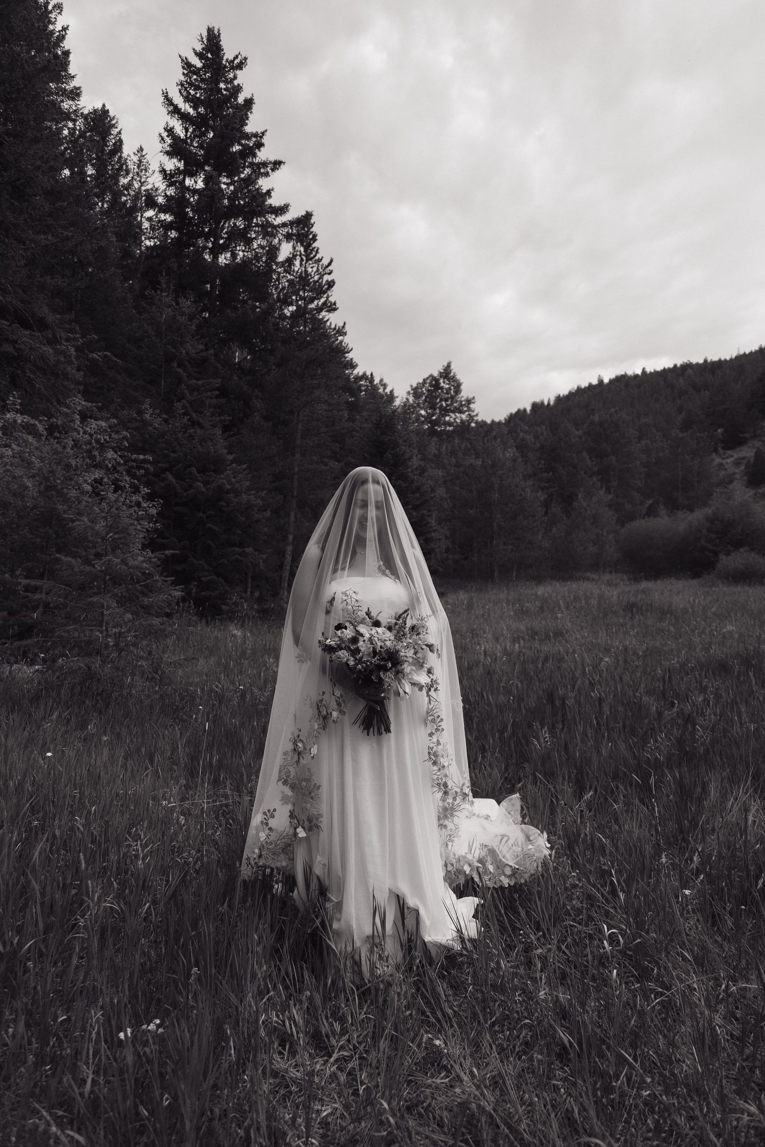 Black and white photo of a bride in a flowing veil and gown holding a bouquet in a forest meadow.