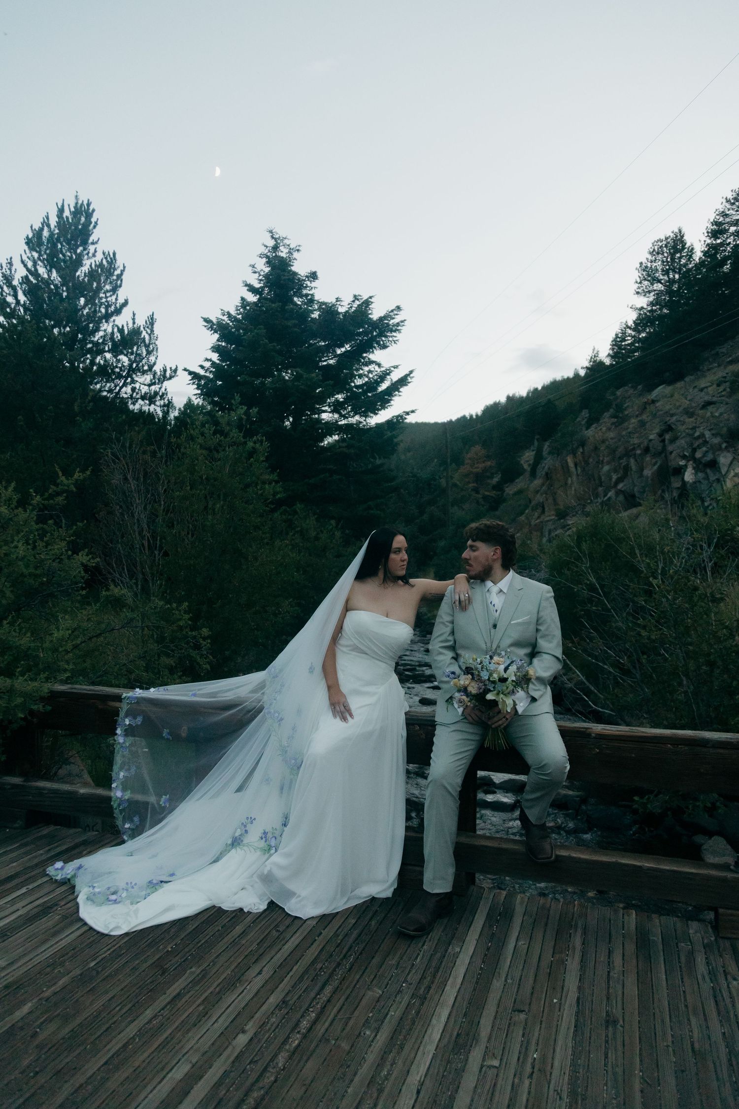 Bride in white gown with flowing veil and groom in gray suit pose on wooden bridge surrounded by trees.