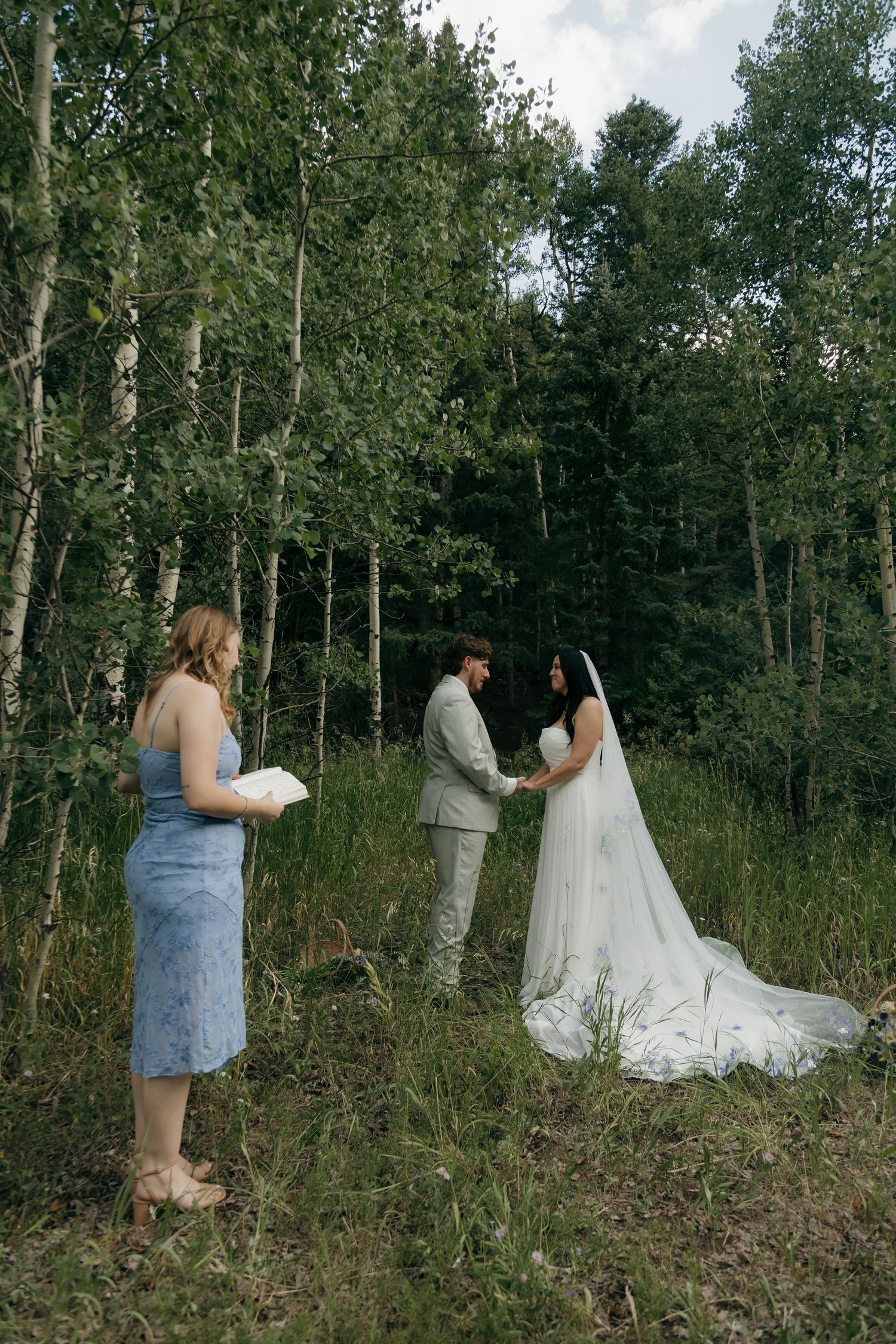 Bride and groom exchange vows in a lush forest clearing with an officiant holding a book during an intimate outdoor wedding ceremony.