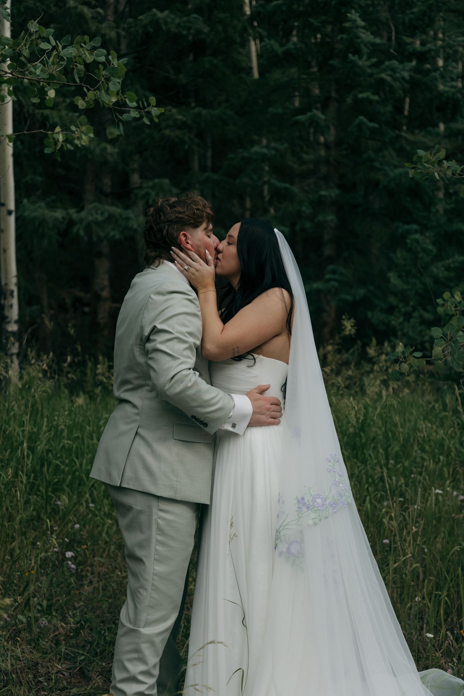 Bride and groom share first kiss surrounded by lush green trees in romantic outdoor wedding.