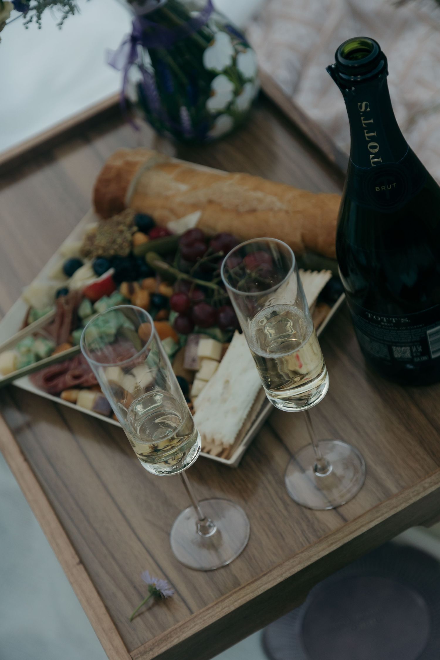 Two champagne flutes beside a charcuterie board and Tott's sparkling wine bottle on a wooden tray.