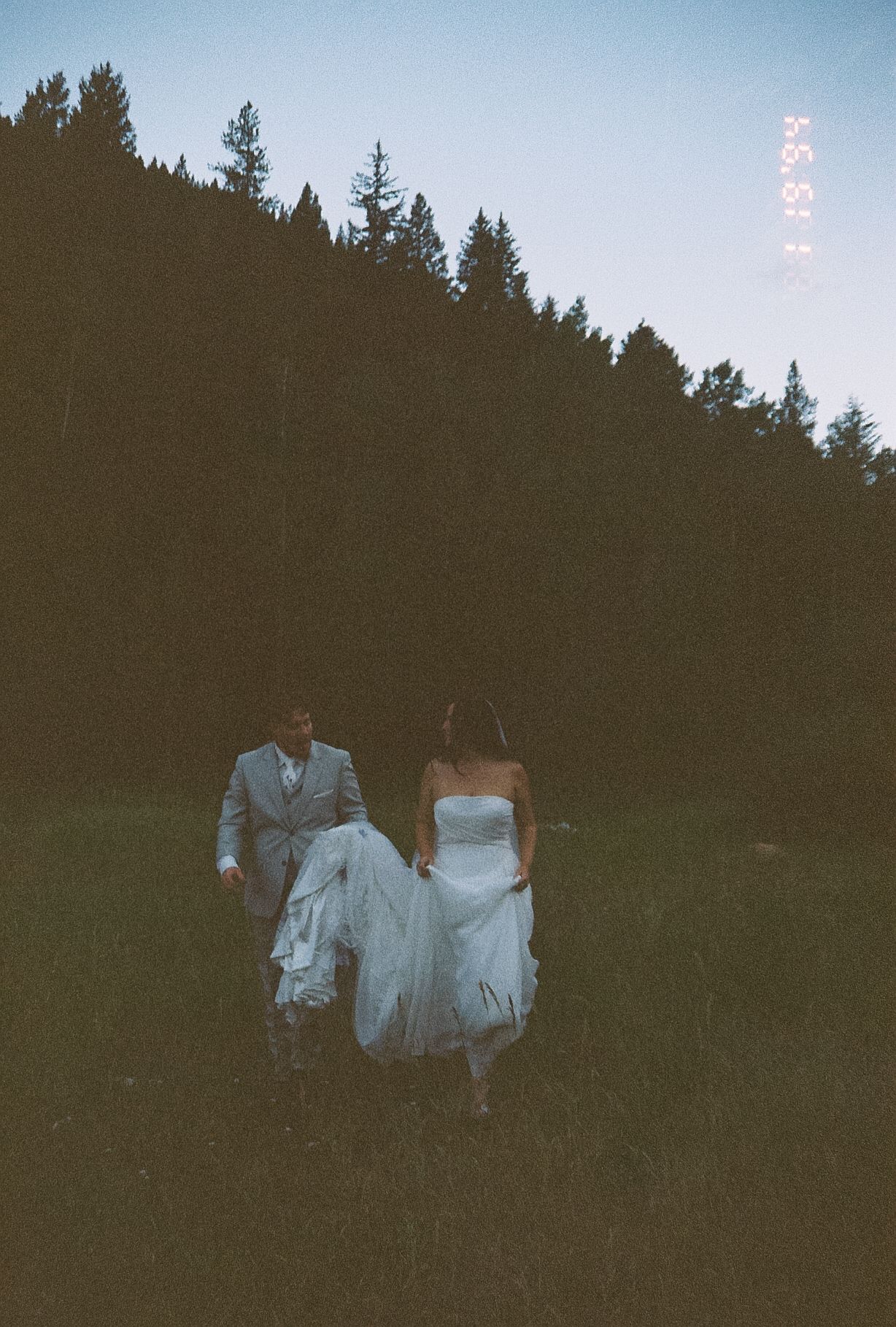 Bride and groom walk through dusky mountain meadow at twilight, groom carrying brides white wedding dress train.